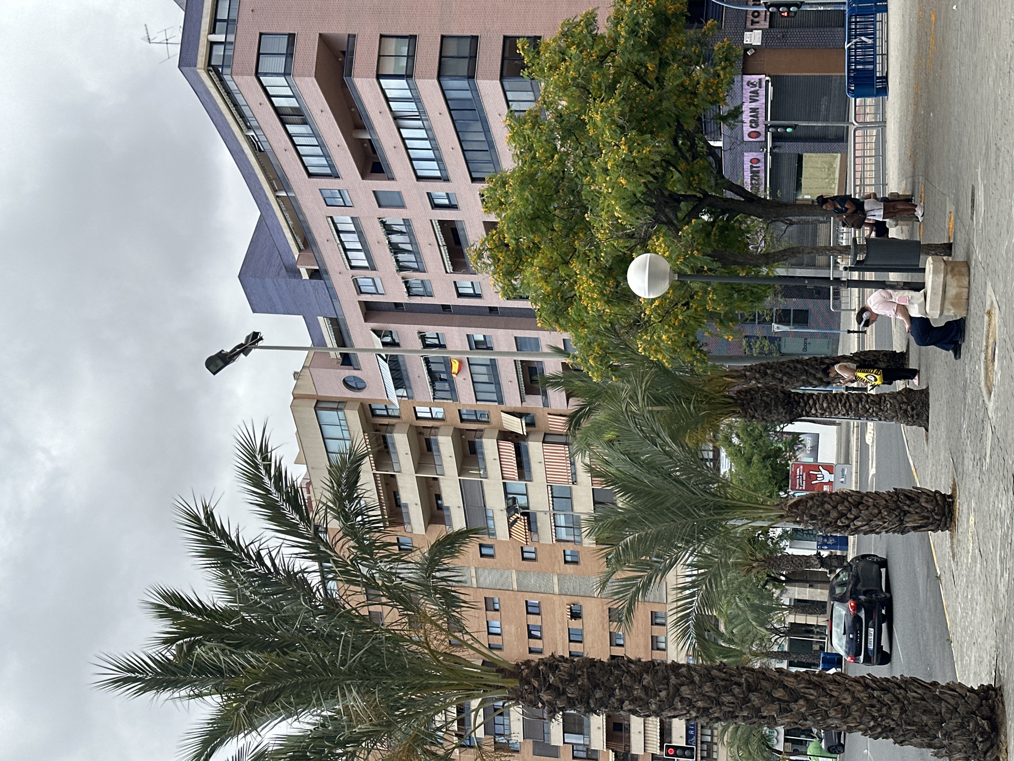 This is a medium-shot, eye-level, outdoor photograph taken on a cloudy day in an urban environment. In the foreground, several large palm trees with thick, textured trunks and feathery fronds dominate the left side of the frame. To the right of the palm trees, a sidewalk is visible, paved with grey interlocking pavers. A few people are present on the sidewalk: one person, wearing headphones and a pink shirt, is sitting on a stone bench, looking at their phone. Another person, wearing a white shirt and shorts, stands near a trash can with a backpack. Further to the right, two more people are standing.
The background features a row of multi-story buildings. The building immediately behind the trees is light brown with visible balconies and windows, some equipped with striped awnings. To the right of this building is a taller, pink-brick building with a curved facade, featuring numerous windows and balconies. A small Spanish flag is visible on a flagpole on the side of one of the buildings. Between the buildings and the sidewalk, a street is visible with a black car driving away from the camera. Several trees with green foliage and yellow flowers are also present on the sidewalk. Streetlights and traffic lights are also visible. The sky is overcast, contributing to the diffused lighting of the scene.