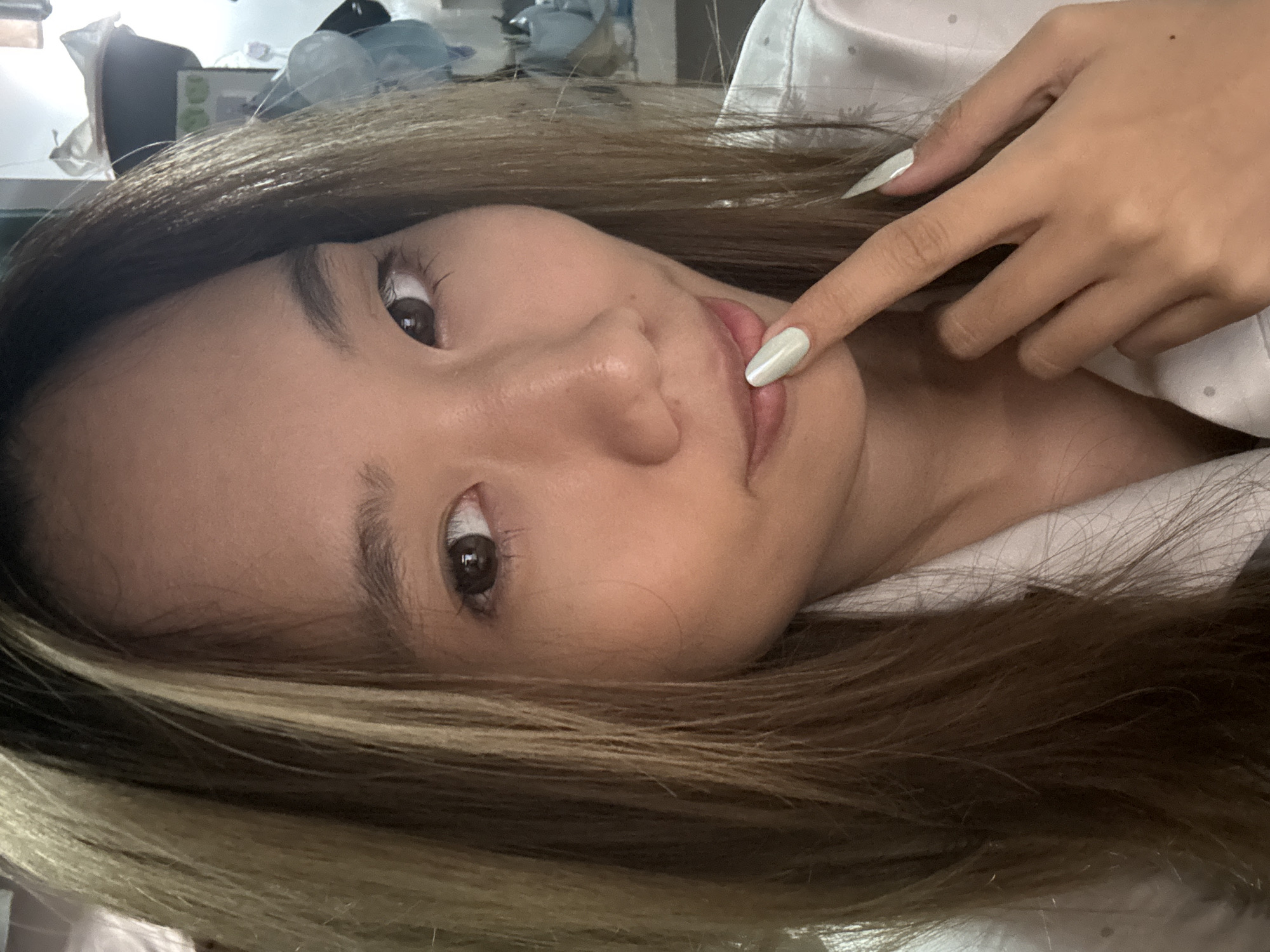 A close-up, tilted view captures a young Asian woman indoors. Her hair, highlighted with blonde streaks, frames her face. She has light skin, brown eyes, and full lips. Her right index finger touches her lower lip, and her nails are painted with a light color. She wears a light-colored, patterned top. The background is blurred, showing glimpses of items on a shelf and a white fabric surface. There is no indication of this image's location in Nan'an District, China.