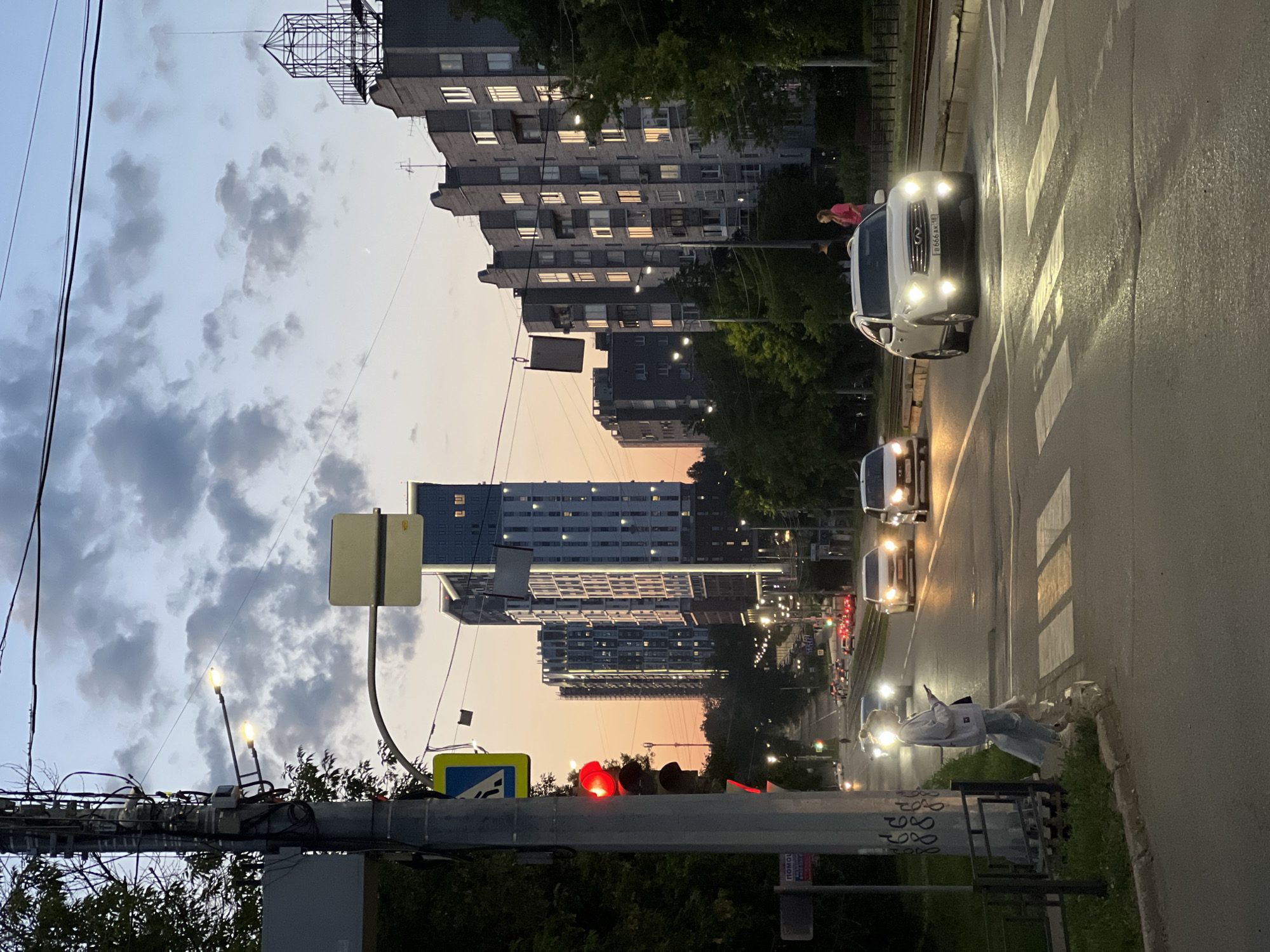 This is a detailed image of an urban street in Izhevsk, Udmurt Republic, Russia, captured at dusk. The scene depicts Proletarskaya Street, looking approximately north, with the prominent "Akvamarin" residential complex visible in the background. The sky shows a gradient from a warm orange-pink hue near the horizon, indicative of sunset, transitioning to a pale blue-grey overhead, with scattered grey clouds. A faint crescent moon is visible in the upper right portion of the sky. In the immediate foreground on the left, a utility pole is heavily laden with numerous black cables, two illuminated streetlights extending from it, and a traffic light showing a solid red signal. Below the traffic light, a blue and white pedestrian crossing sign is attached. Further down the pole, a partially visible sign reads "ПОМОЩЬ" (Help) and "ТЕЛ." (Tel.) with graffiti "888" beneath. Adjacent to the pole, a young woman in a light-colored sweatshirt and blue jeans stands on the curb, looking at a smartphone, her face illuminated by reflections from car headlights as she waits to cross a zebra pedestrian crossing. A low black metal fence runs along the sidewalk beside her. The asphalt road features a clearly marked zebra crossing in the midground, with illuminated yellow paint reflecting light. Several vehicles are present on the road. A white Infiniti SUV with its headlights and fog lights illuminated is positioned centrally, displaying Russian license plate "B 666 AK 18". Behind it, another white car with illuminated headlights is visible. In the distance, numerous cars are seen with their headlights and taillights on, moving along the street. Tram tracks run parallel to the road on the right side of the frame. Streetlights are visible lining both sides of the road, with many actively illuminated. On the right side of the street, an older, multi-story residential building constructed of grey brick features numerous windows, many of which are illuminated, reflecting the evening light. A metal, cage-like structure is visible on its rooftop. Further down the sidewalk near this building, two pedestrians, a man and a woman in a pink top, are standing. Dominating the background are several modern high-rise residential towers, characteristic of the "Akvamarin" complex (located at the intersection of Proletarskaya Street and Kirova Street). These buildings are slender, with light-colored facades and distinct vertical strip lighting along their sides, many windows also illuminated. Behind these, other less distinct buildings are visible, receding into the