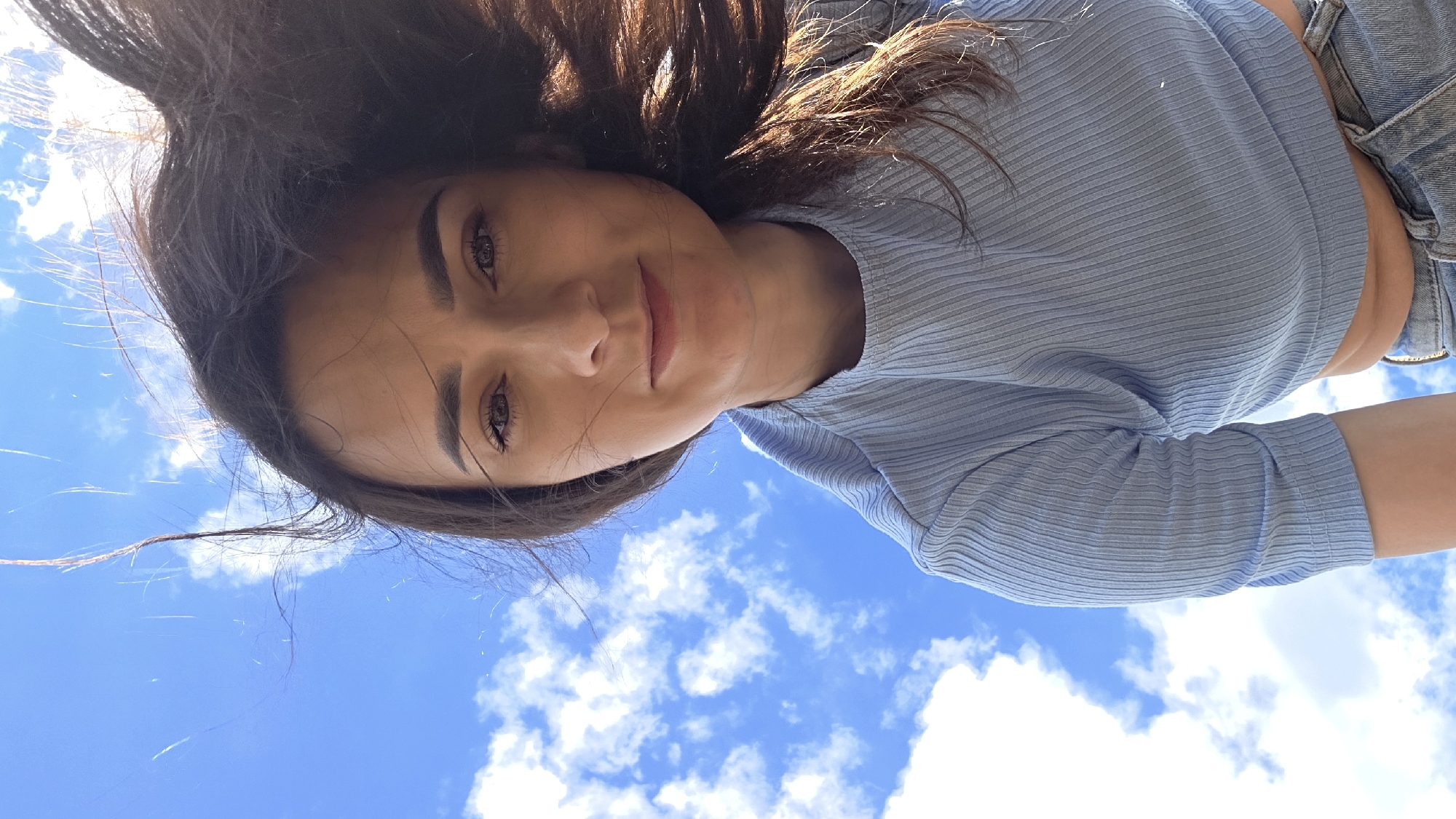 A low-angle, medium shot captures a young woman from the chest up, positioned against a bright blue sky filled with white clouds. Her long, dark hair is windswept, with strands flying across her face and into the frame from the right. She has medium-toned skin, dark eyes, and subtle makeup. She is wearing a light blue, ribbed, short-sleeved shirt and blue denim jeans. The camera appears to be looking up at her. The location is Дарьино, Russia.