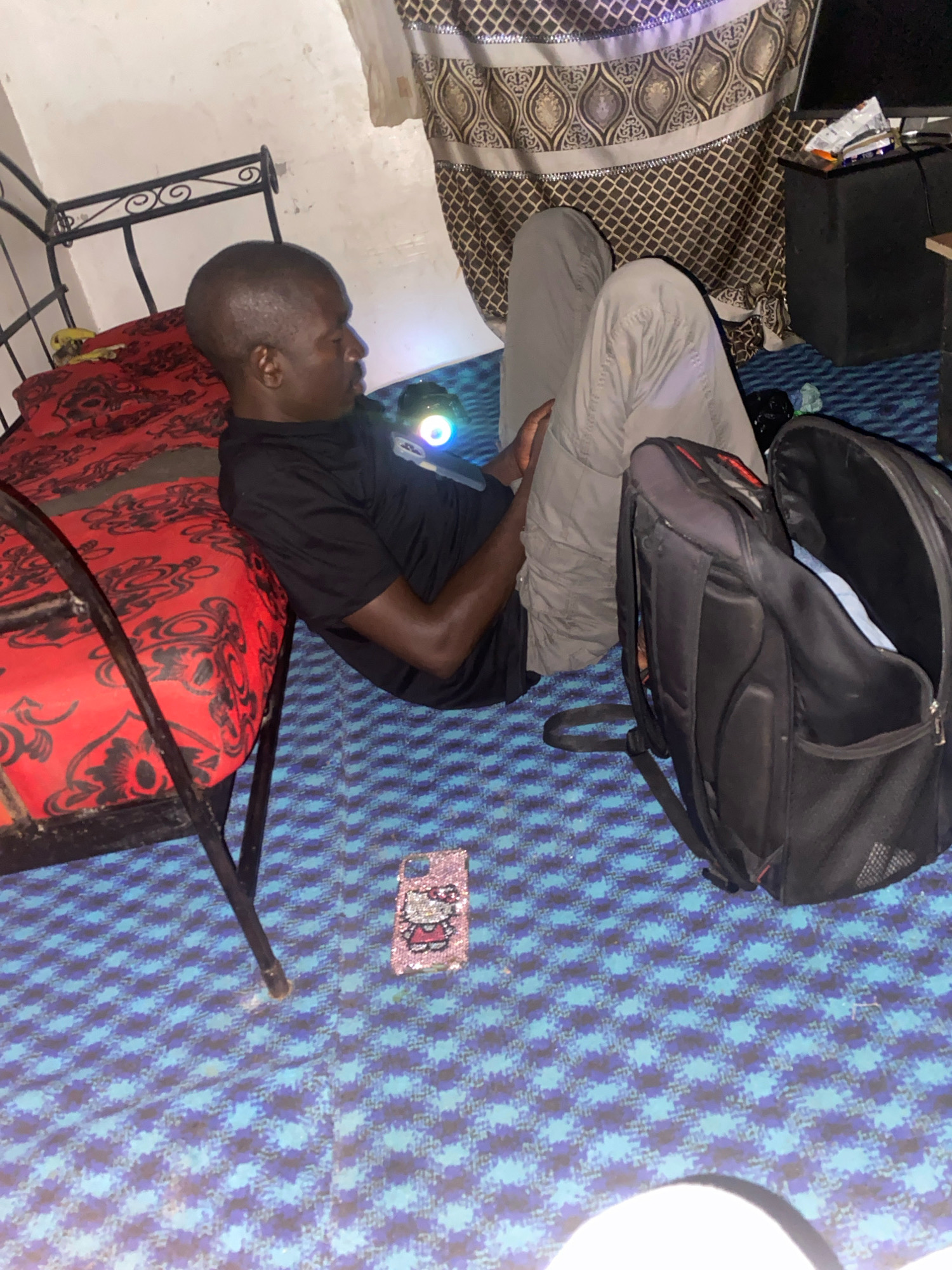 In a dimly lit room, a dark-skinned individual with a shaved head is reclining on the floor. They are wearing a black t-shirt and light gray pants. To their left is a red and black patterned mattress on a bed frame. In front of them, on a blue and black patterned floor covering, is a pink Hello Kitty phone case covered in rhinestones and a partially opened black backpack. The background shows a patterned curtain and what appears to be a television screen on a black cabinet. The overall impression is of a modest living space, possibly a bedroom, within a residential setting in Jos, Nigeria.