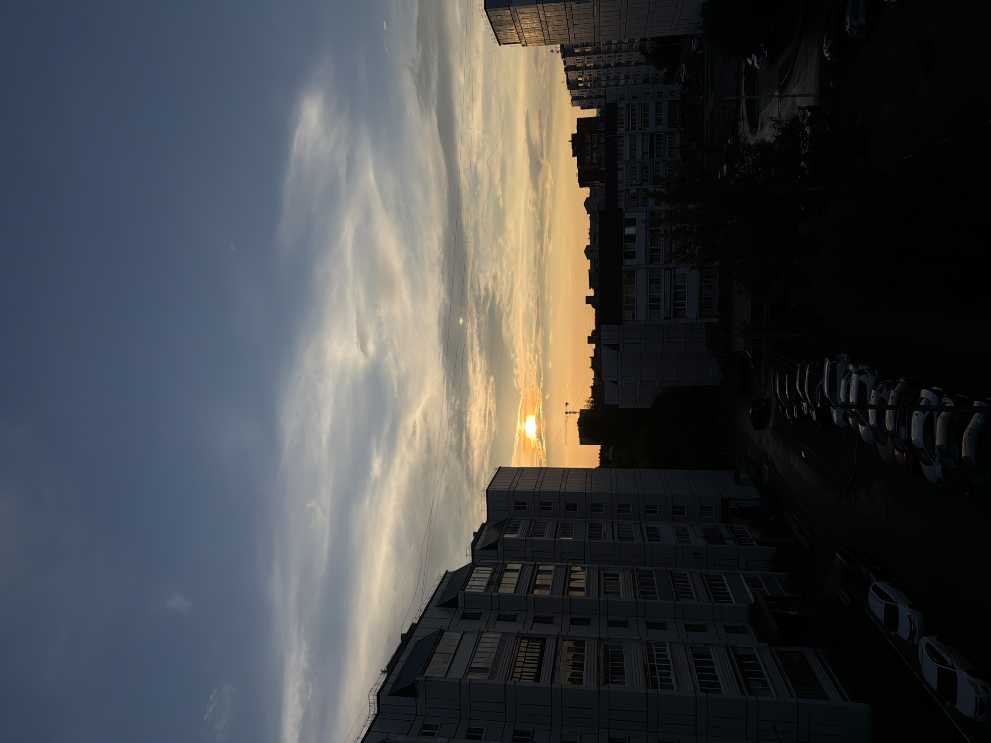 The image depicts a view of an urban area in Chaikovsky, Russia, during sunset. The sky is filled with a mix of blue and orange hues, with scattered clouds. The sun is visible, low on the horizon, casting a warm glow.
In the foreground, several multi-story residential buildings, typical of Soviet-era architecture, are silhouetted against the twilight sky. Their facades are a pale grey, with numerous windows visible. Electrical wires are strung between some of the buildings.
Below the buildings, a street is visible, lined with parked cars. The lighting conditions indicate it is either early evening or late afternoon, with shadows lengthening. The environment appears to be a residential district. No individuals are clearly discernible in the image.
