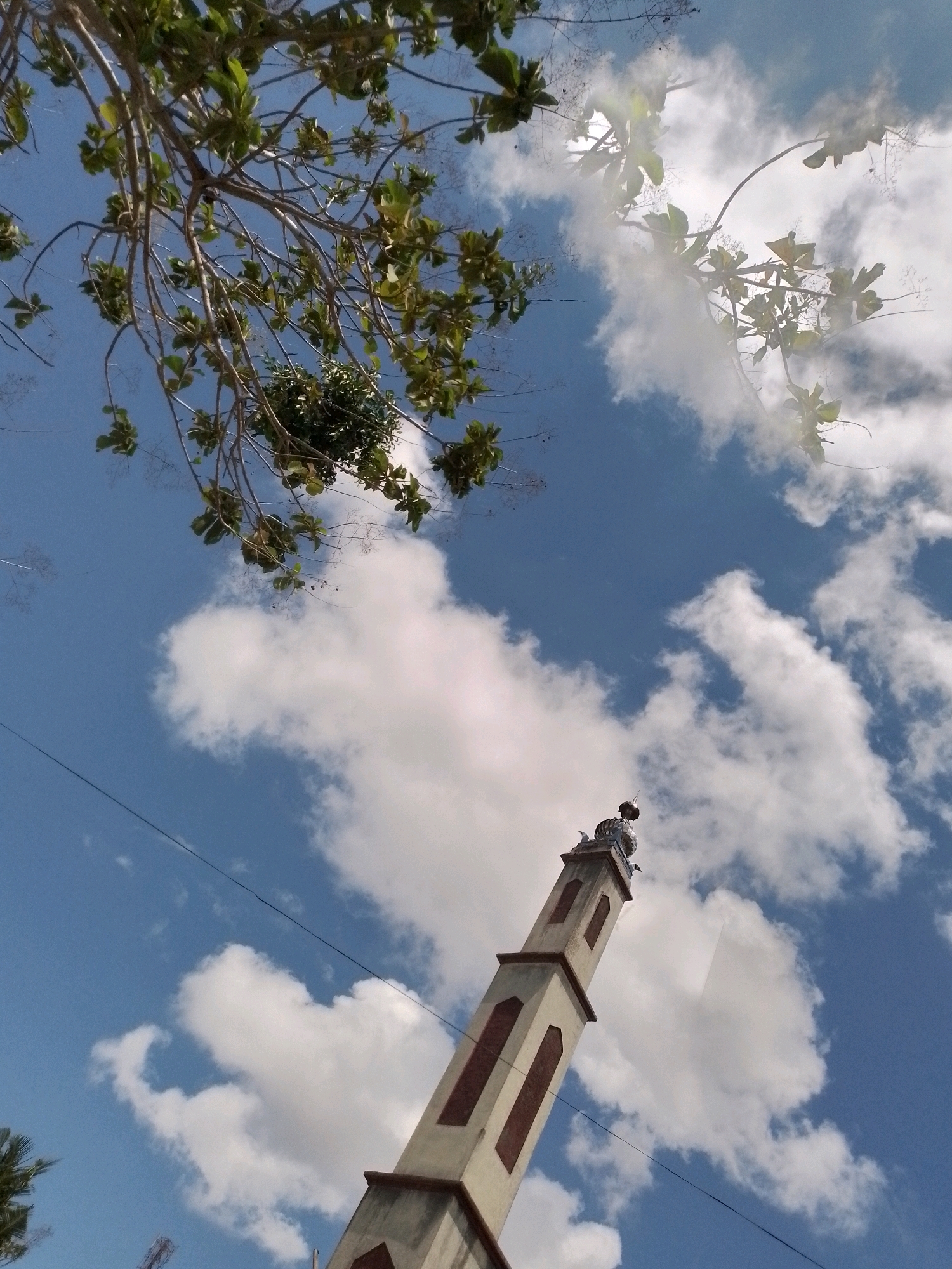 An upward shot shows a minaret from a mosque in Wonosari, Indonesia, against a bright blue sky with white clouds. The minaret, a tall, slender structure, is light-colored with rectangular window openings. At the top of the minaret is a decorative finial. To the left, tree branches with green leaves hang into the frame, obscuring parts of the sky. The overall scene suggests a sunny day with a few clouds.