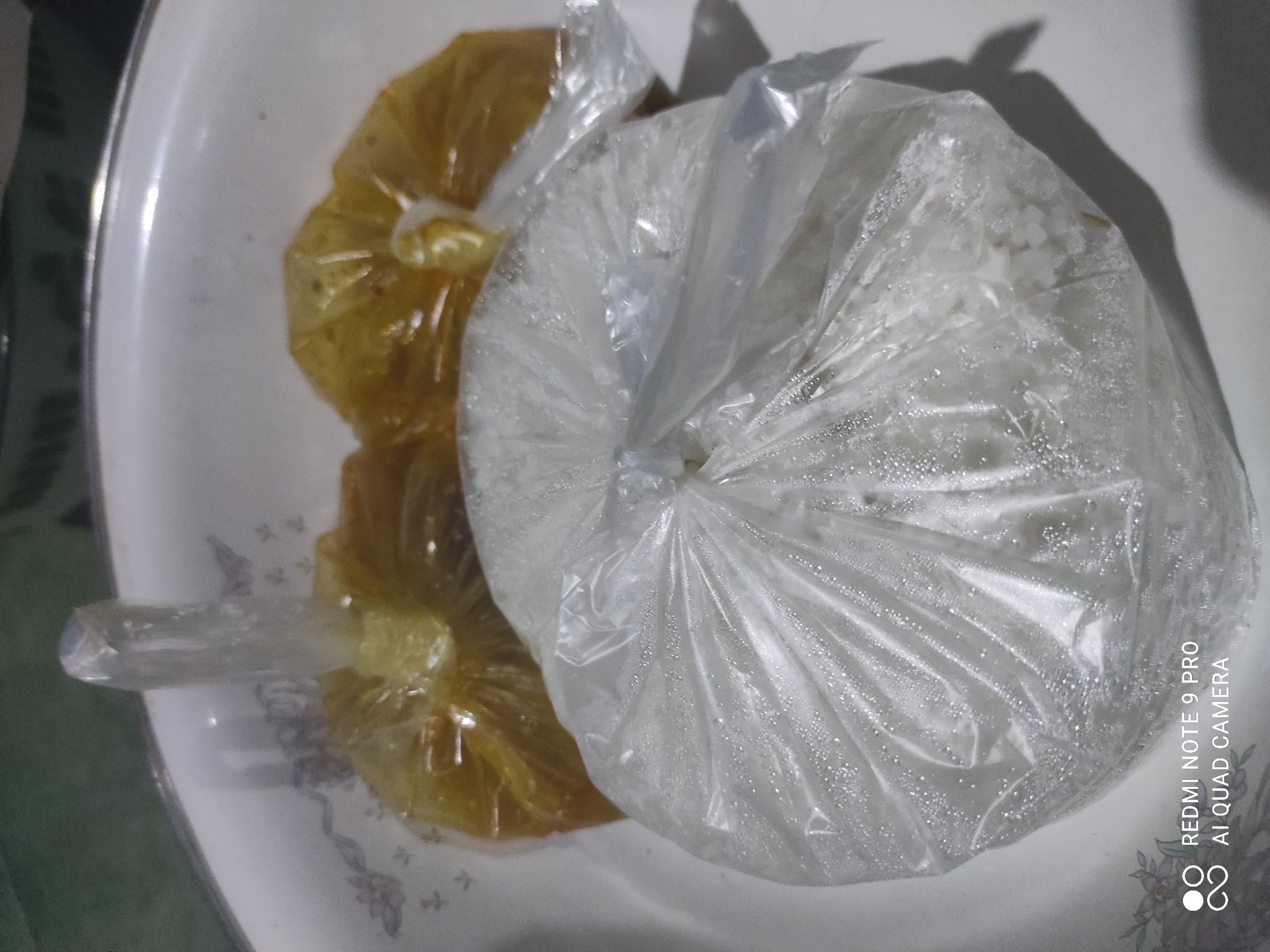 In the city of Sakra, Indonesia, a white ceramic bowl contains three servings of food, each enclosed in clear plastic packaging. One serving, in the foreground, is white rice. To the left and behind the rice, two smaller servings contain a yellowish, gravy-like substance. The scene is lit from above, casting shadows to the right of the food items. The overall impression is of a prepared meal, possibly for takeaway or delivery.