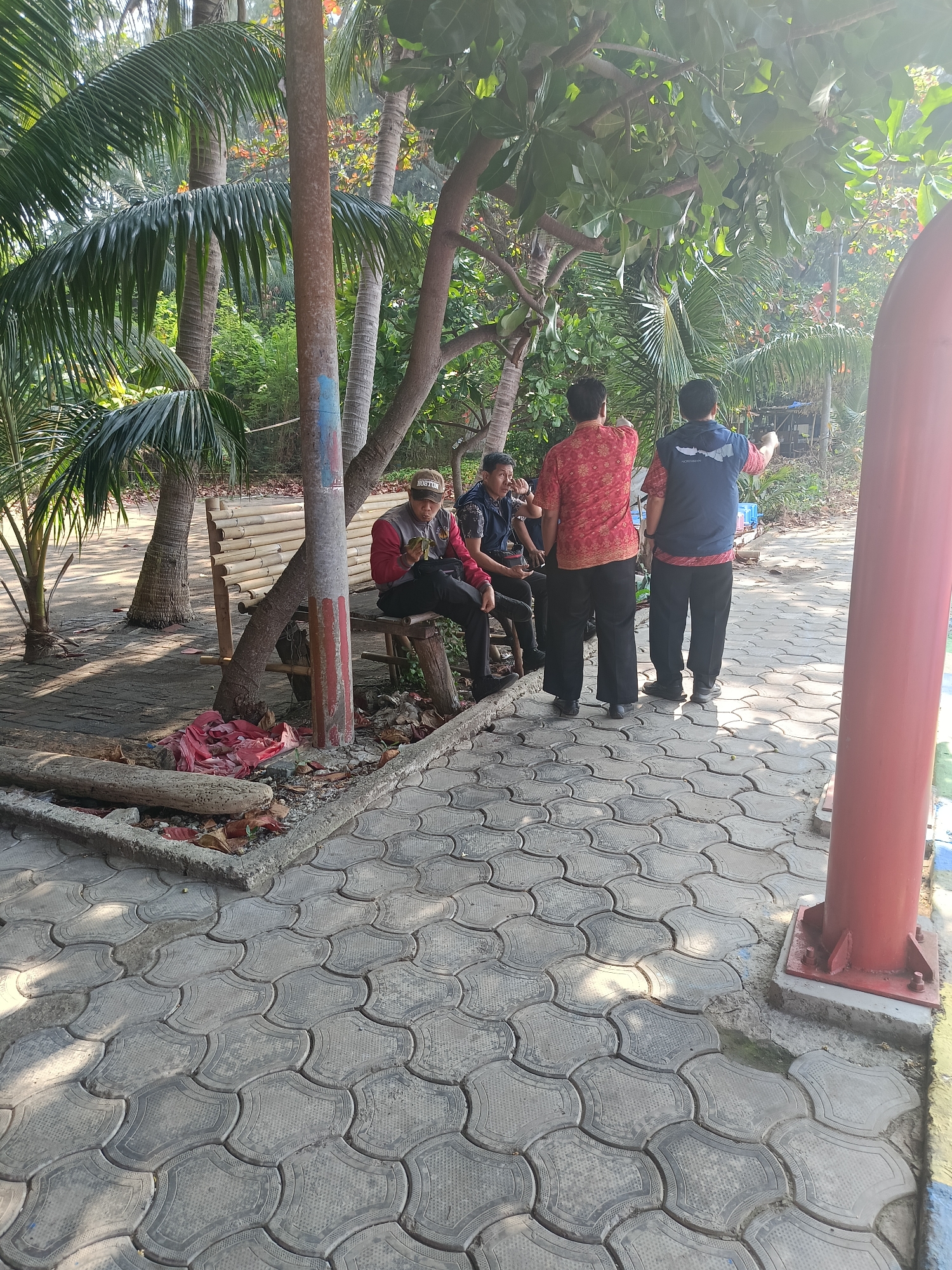 The image depicts a group of men gathered outdoors near a park area. In the foreground, a patterned paved walkway leads towards a red cylindrical structure. To the left of the walkway, a person is seated on a wooden bench made of bamboo. Another man, wearing a baseball cap and a red long-sleeved shirt, is also seated on the bench and appears to be eating. Behind him, two other men stand, one wearing a red and black patterned shirt and the other a navy blue vest over a red shirt. Both standing men are gesturing with their hands, possibly indicating something in the distance or engaged in conversation. The environment is lush with palm trees and other vegetation, suggesting a tropical or subtropical climate, common in the Special Capital Region of Jakarta, Indonesia. The lighting suggests it is daytime, with sunlight casting shadows on the walkway and vegetation. There are fallen leaves and debris on the ground near the bench.