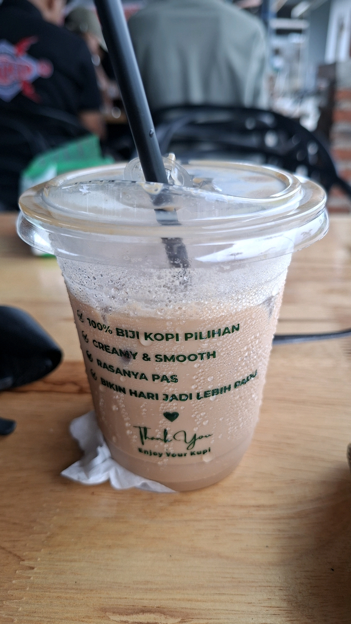 A close-up, eye-level, vertical shot focuses on a transparent plastic cup of iced coffee. The cup is condensation-covered and sits on a wooden table. Green text on the cup reads "100% BIJI KOPI PILIHAN" (100% Selected Coffee Beans), "CREAMY & SMOOTH", "RASANYA PAS" (Taste is Right), and "BIKIN HARI JADI LEBIH BAIK" (Makes the Day Better). Below this, a small green heart is visible, followed by "Thank You" in script and "Enjoy Your Kupi" in smaller print. A black straw extends from the clear plastic lid, which is slightly askew. In the blurred background, a person wearing a green shirt is visible, and further back, another person is partially visible in a black shirt with a red graphic. The overall setting appears to be an indoor cafe with a wooden interior. This image was captured in Keudebayu, Indonesia.
