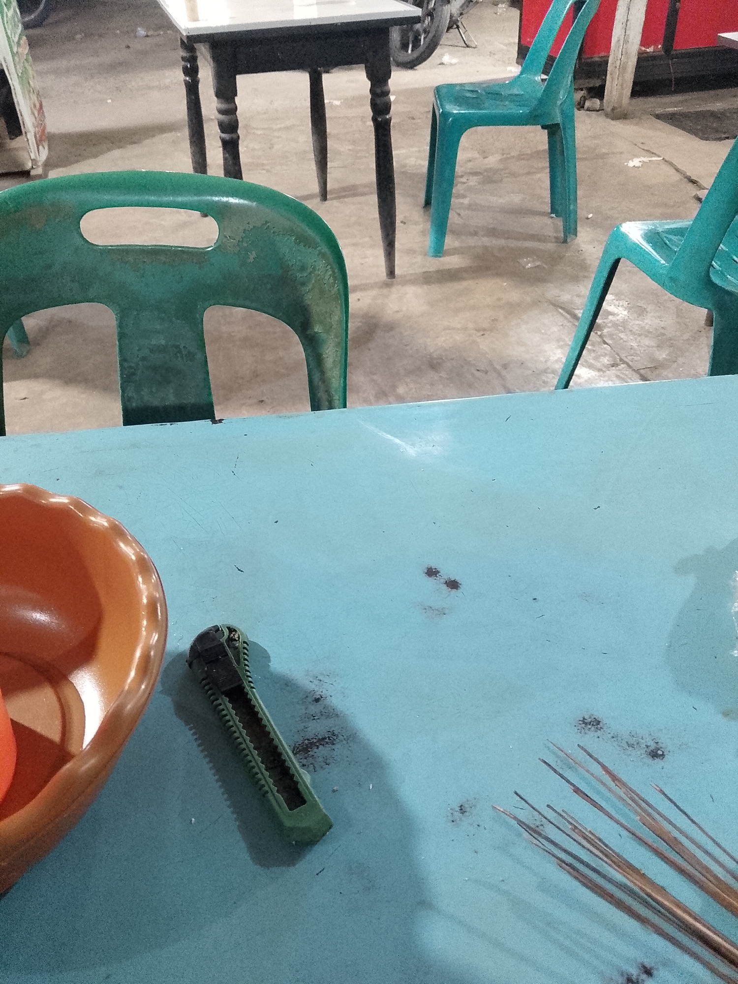 The image captures an outdoor dining area, possibly in Lhokseumawe, Indonesia, given the prompt context. The focus is a turquoise-colored table, on which are an orange bowl, a green utility knife housing, and several thin, brown sticks or skewers. The table surface also exhibits minor stains and debris. In the background, there are green plastic chairs and a black table with white top, indicating a typical outdoor eatery setting. A glimpse of a motorcycle wheel is visible further back, suggesting a street or open area. No people are visible in the frame. The setting conveys a casual, perhaps even somewhat utilitarian atmosphere.