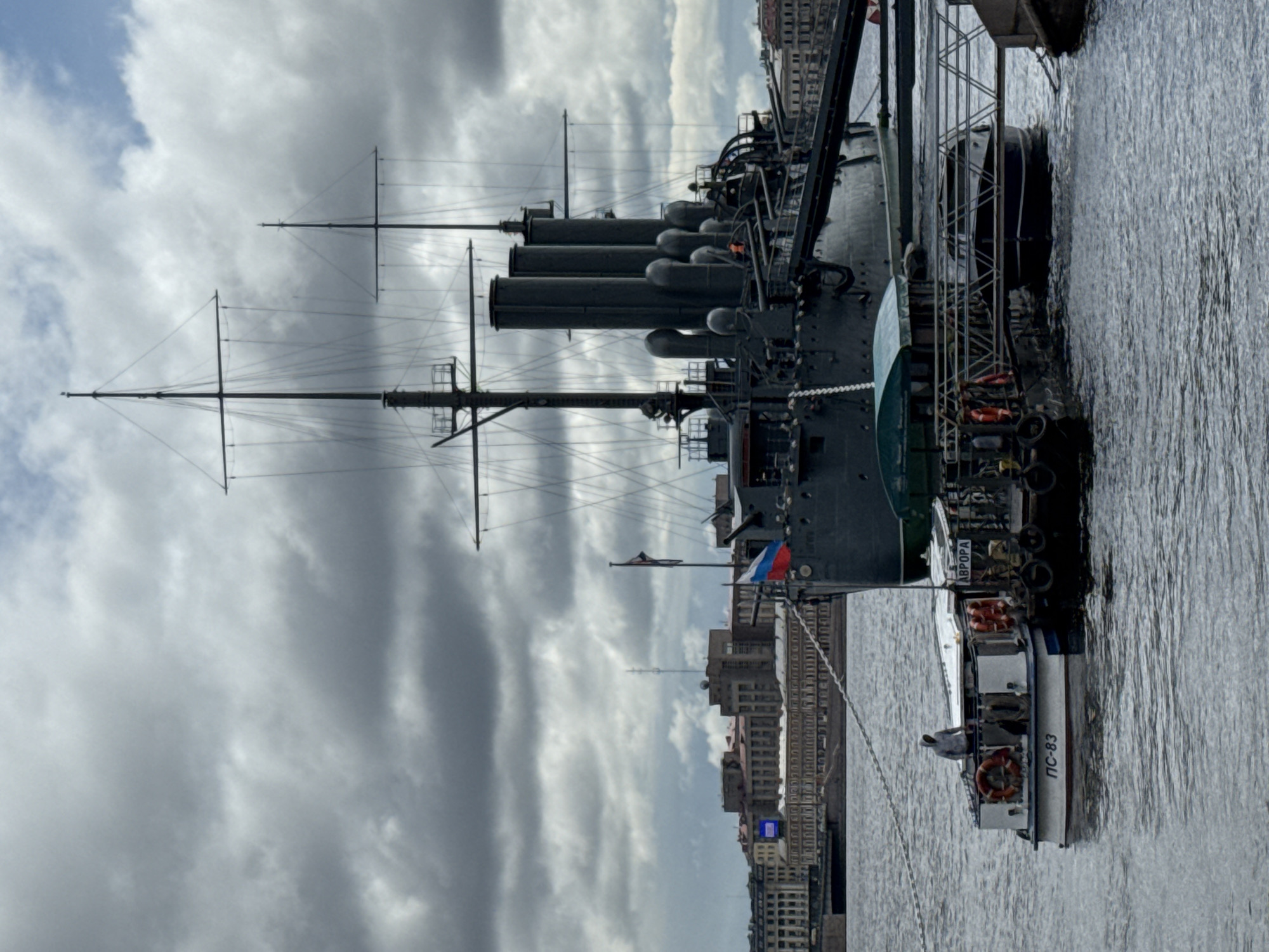 Here's a description of the image:
The image showcases the Cruiser Aurora, a historic Russian warship, docked in Saint Petersburg, Russia. The vessel is positioned adjacent to a quay along the Neva River, evident by the urban landscape and architectural features of the city along the embankment in the background.
The ship's black hull, multiple funnels, and rigging are prominently displayed. A smaller white boat, labeled "ПС-83," is moored alongside the Aurora, and there is a person visible on the small boat.
The sky overhead is overcast with textured gray clouds. The water appears choppy with minor wave action. The Russian flag is visible flying from the Aurora. The overall scene is viewed from a medium distance, capturing the boat and the general river and sky backdrop.