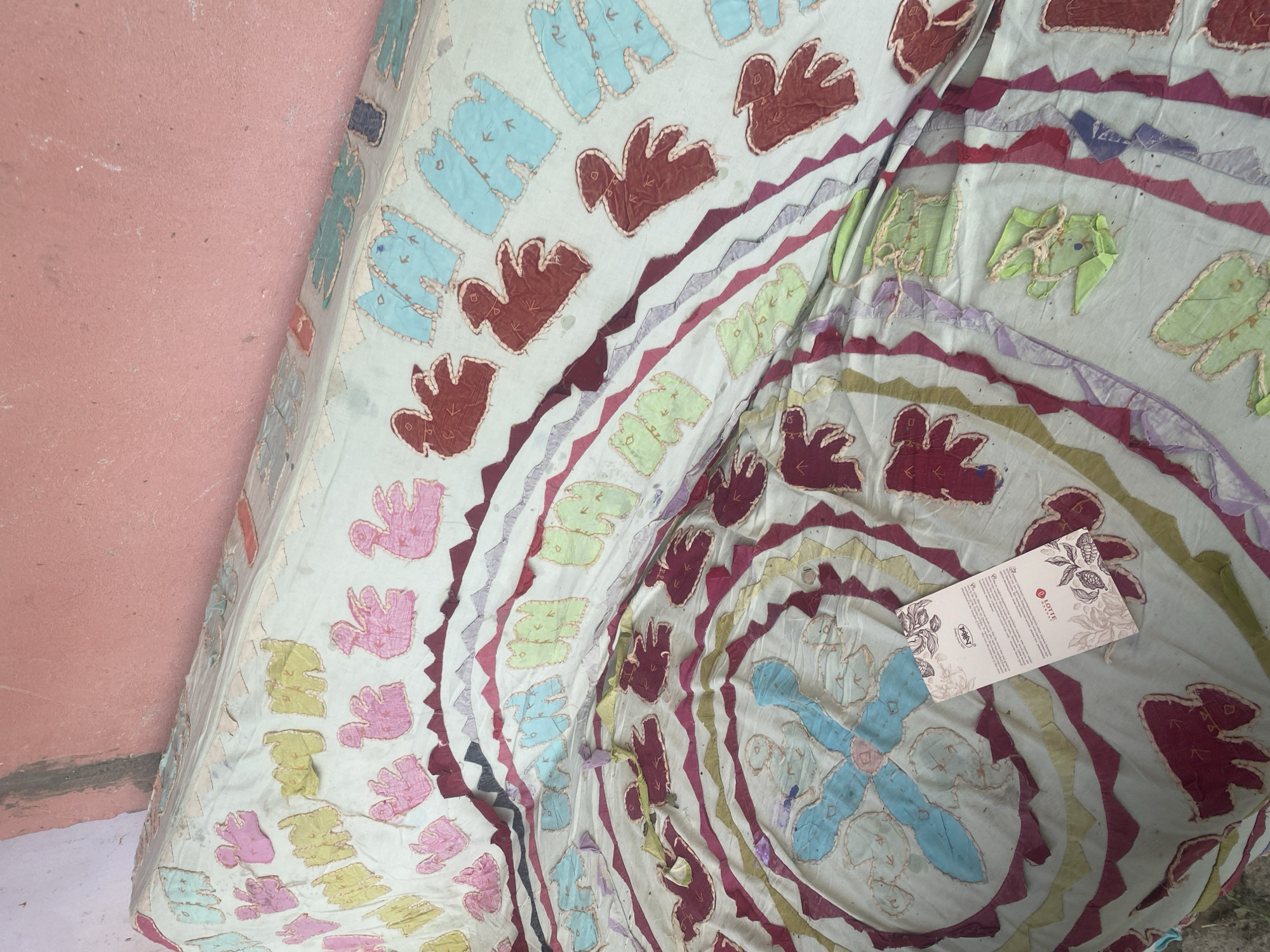 The image shows a close-up of a textile with a colorful, patterned design, likely a quilt or blanket. The textile is laid out against a pink, textured wall and a light-colored floor. The design features a variety of shapes and colors, including what appear to be stylized birds or animals in shades of blue, red, pink, and green, stitched onto a light-colored fabric. There are also concentric circular patterns with zig-zagging lines and diamond shapes in burgundy, purple, and yellow. A small white tag with text and floral designs is placed on the textile. The tag has text that reads "LOTTE" in red lettering, and "PARRY" above a logo. The lighting suggests it is daytime, with no specific weather conditions evident. The setting appears to be indoors or in a sheltered outdoor space.
