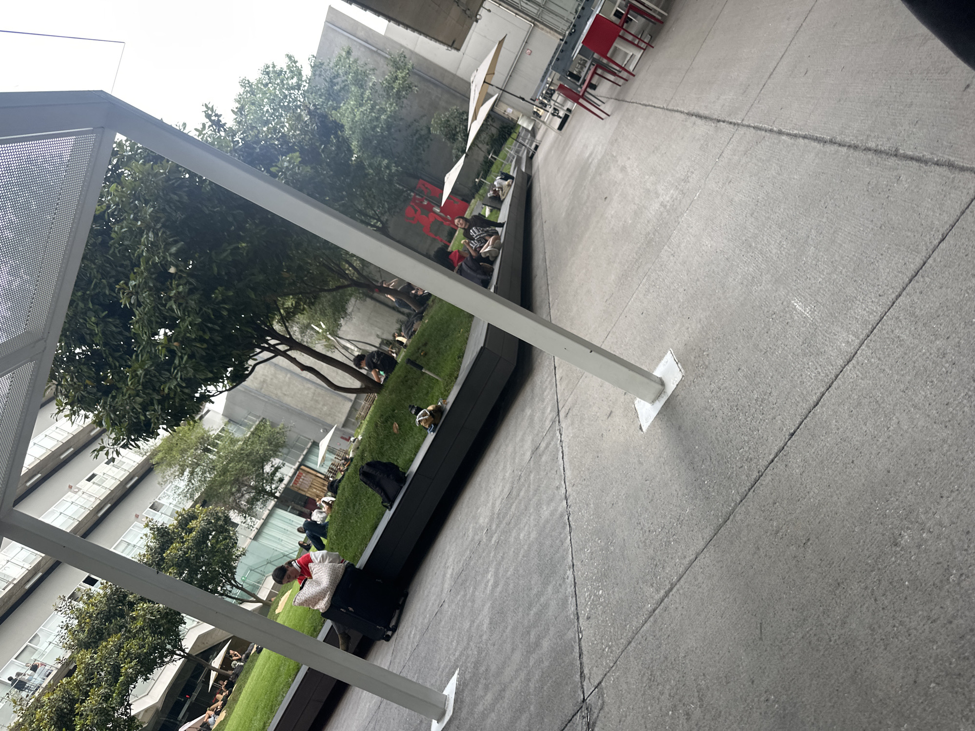 The image depicts an outdoor public space in Mexico City, Mexico. The scene is partially covered by a modern architectural structure with a perforated metal roof and white support beams. The area is paved with concrete.
A long, raised concrete planter runs through the center of the frame. This planter contains grass, trees, and various people. Some people are sitting or lying on the planter, relaxing or using electronic devices. One person is seen with luggage, wearing a red and white shirt.
In the background, there is a multi-story building with numerous windows and trees. In the upper right of the image, tables and chairs are visible. The overall scene suggests a relaxed, outdoor environment in an urban setting.