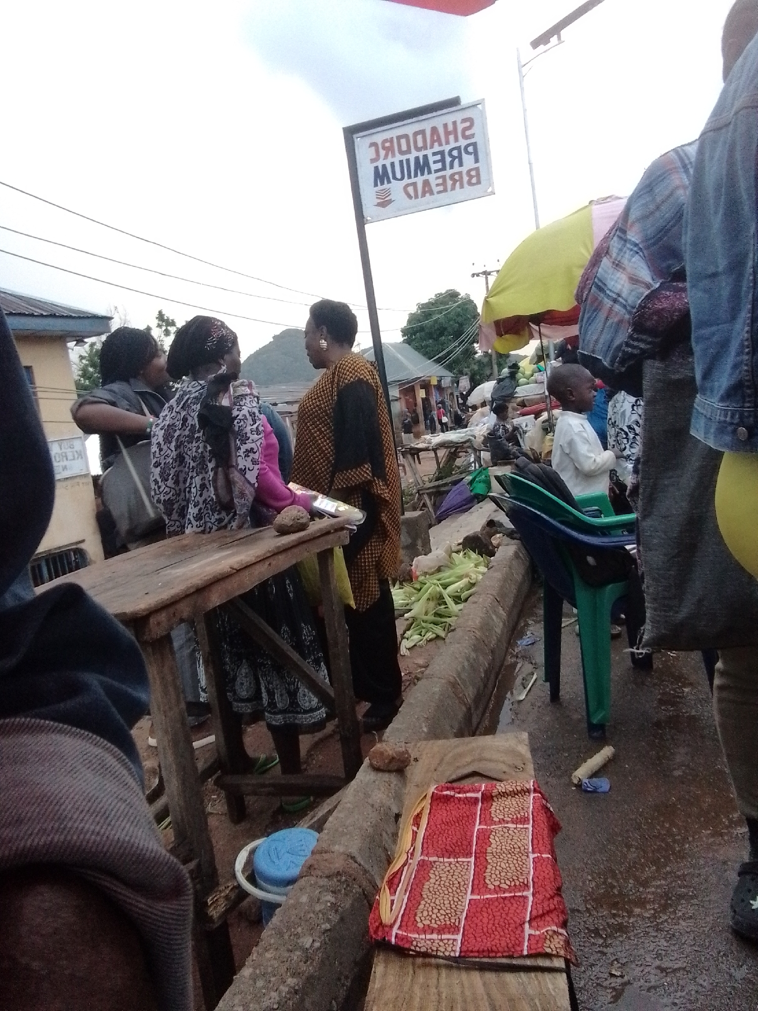 This media file captures a bustling market scene in Gwong, Nigeria. The foreground features a rustic wooden stall laden with what appears to be food items. A colorful red and patterned cloth is draped over a wooden surface, possibly a mat or a bundle of goods, near a blue bucket. Behind the stall, a young boy in a white shirt stands looking to the right, while several women are engaged in conversation. One woman wears a patterned black and white outfit, another a vibrant orange and yellow patterned top, and a third a dark top. The women are positioned around the stall, suggesting they might be vendors or customers.
In the background, a white sign with dark lettering is visible, displaying the text "SHADDC RE" and "MUIMESB" and "BREVD", suggesting it's an advertisement for bread. The market appears to be outdoors, under an overcast sky, indicating it could be morning or afternoon. The ground is wet, suggesting recent rain or damp conditions. The overall atmosphere seems active and social, with people interacting and conducting business. The presence of overhead wires and structures suggests an urban or semi-urban market setting. The clothing worn by the individuals suggests a warm climate.