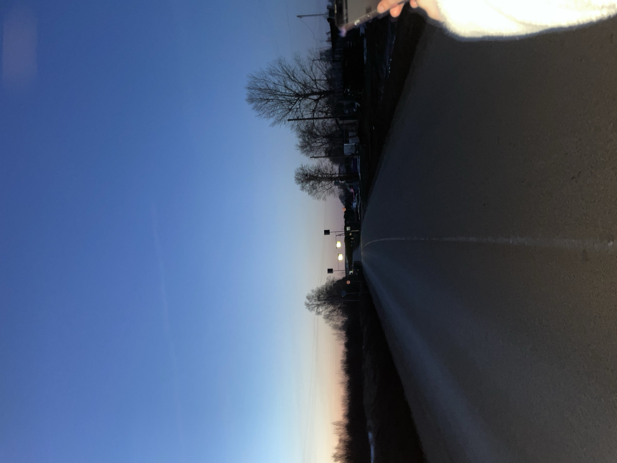 The image shows a road receding into the distance under a gradient sky transitioning from blue to a lighter orange hue on the horizon. The road appears to be paved, with a dividing line visible. Bare trees line the side of the road, suggesting a rural or suburban setting. Streetlights are sparsely placed along the road. A person's hand and part of their white clothing are visible in the upper right corner of the image. The scene's orientation implies it was captured in portrait mode. The lighting indicates it is either dusk or dawn. I cannot confirm if this is Фоки, Russia, without more information.