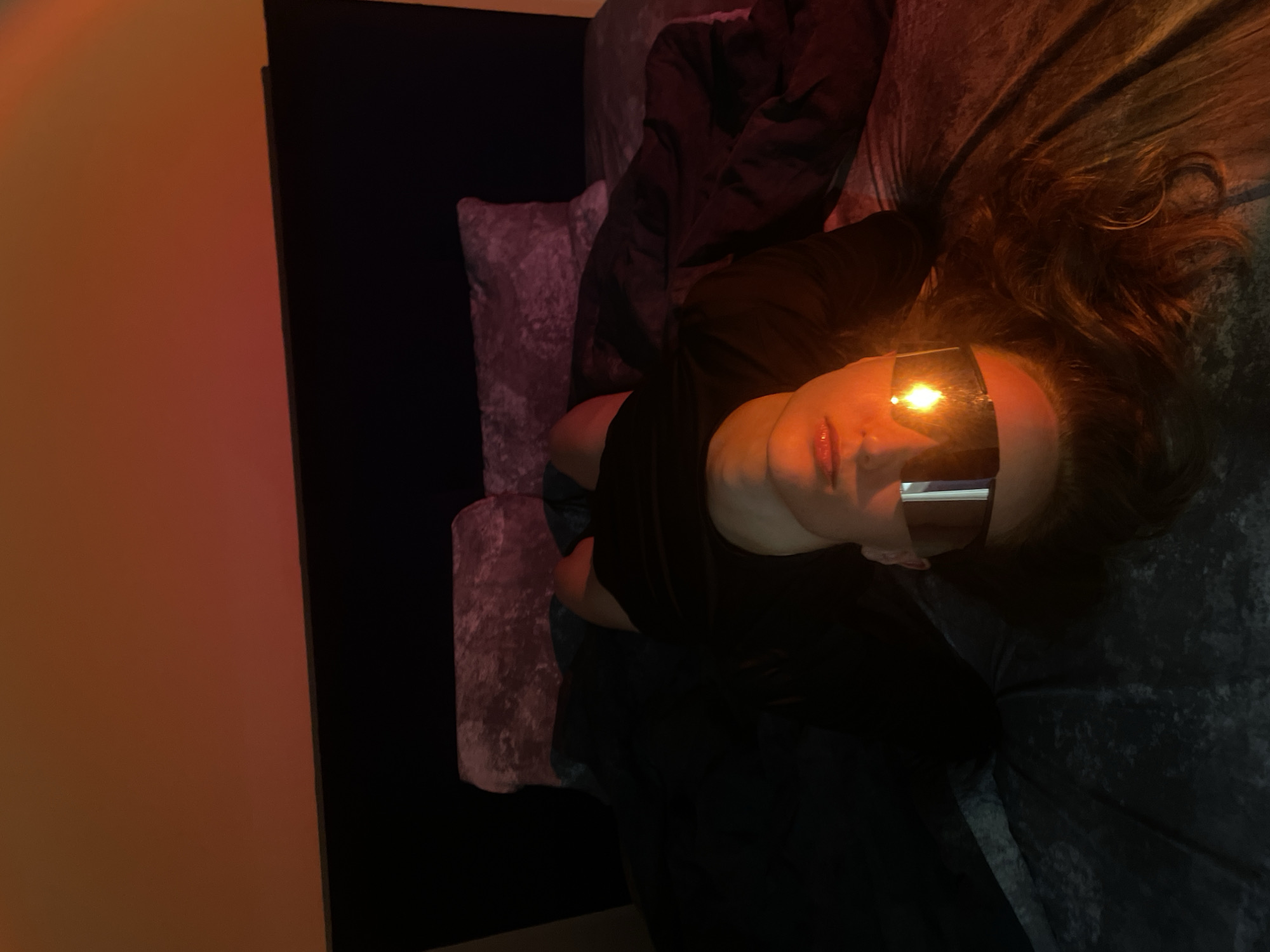 A person lies on a crushed velvet surface of unknown color. They wear dark-tinted eyewear that reflects light. The person has long, wavy hair. The setting appears to be indoors with a wall visible, colored orange on one side and black on the other. The location in the city of Unknown, Unknown is unknown.