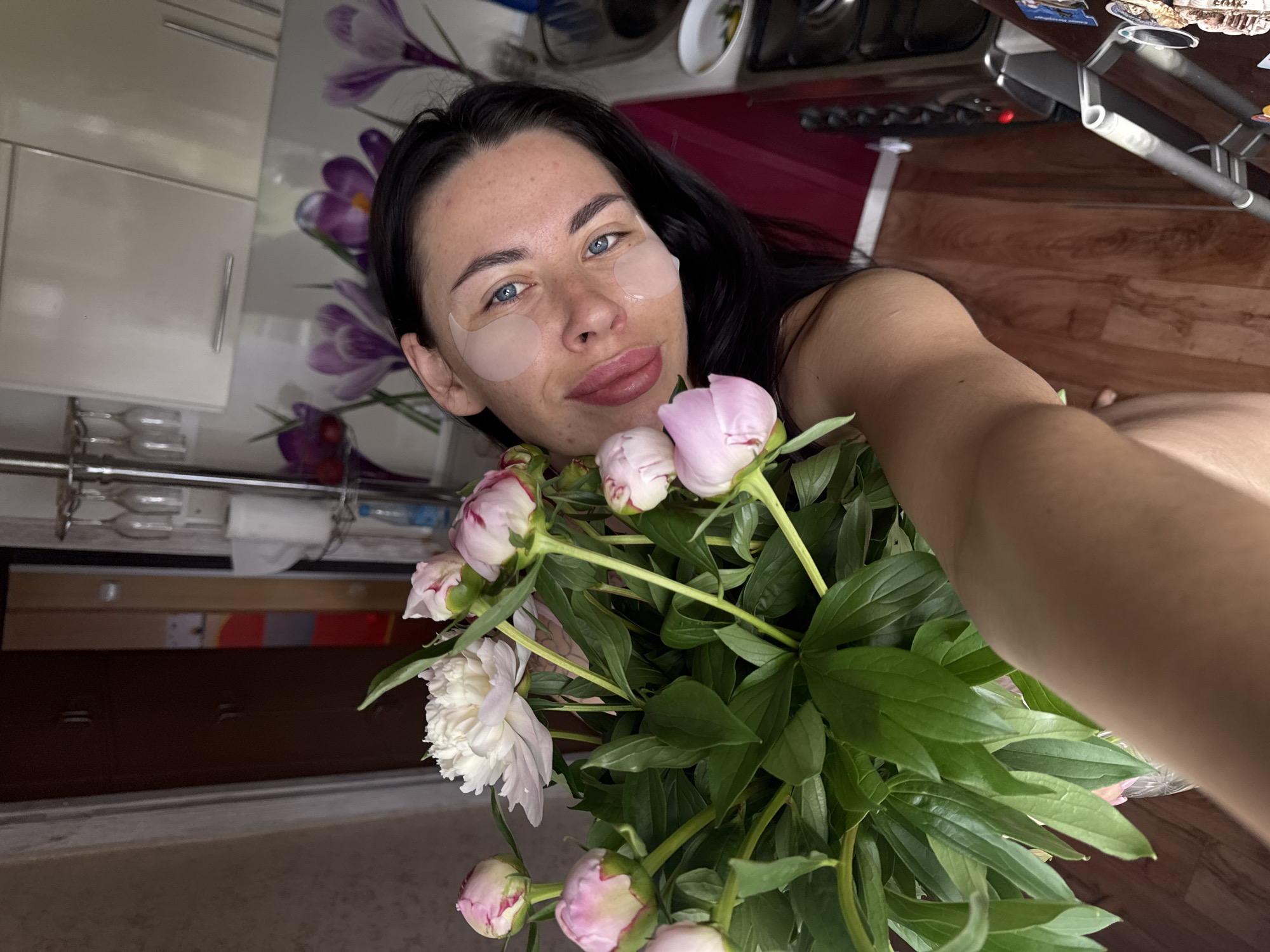A woman with dark hair and blue eyes, wearing eye patches and holding a bouquet of pink and white peonies, is pictured in what appears to be a kitchen. The background shows kitchen cabinets, a floral-patterned backsplash, and a wooden floor. A glimpse of a doorway and a wardrobe is visible to the left. The setting is consistent with an apartment in Chaikovsky, Russia.