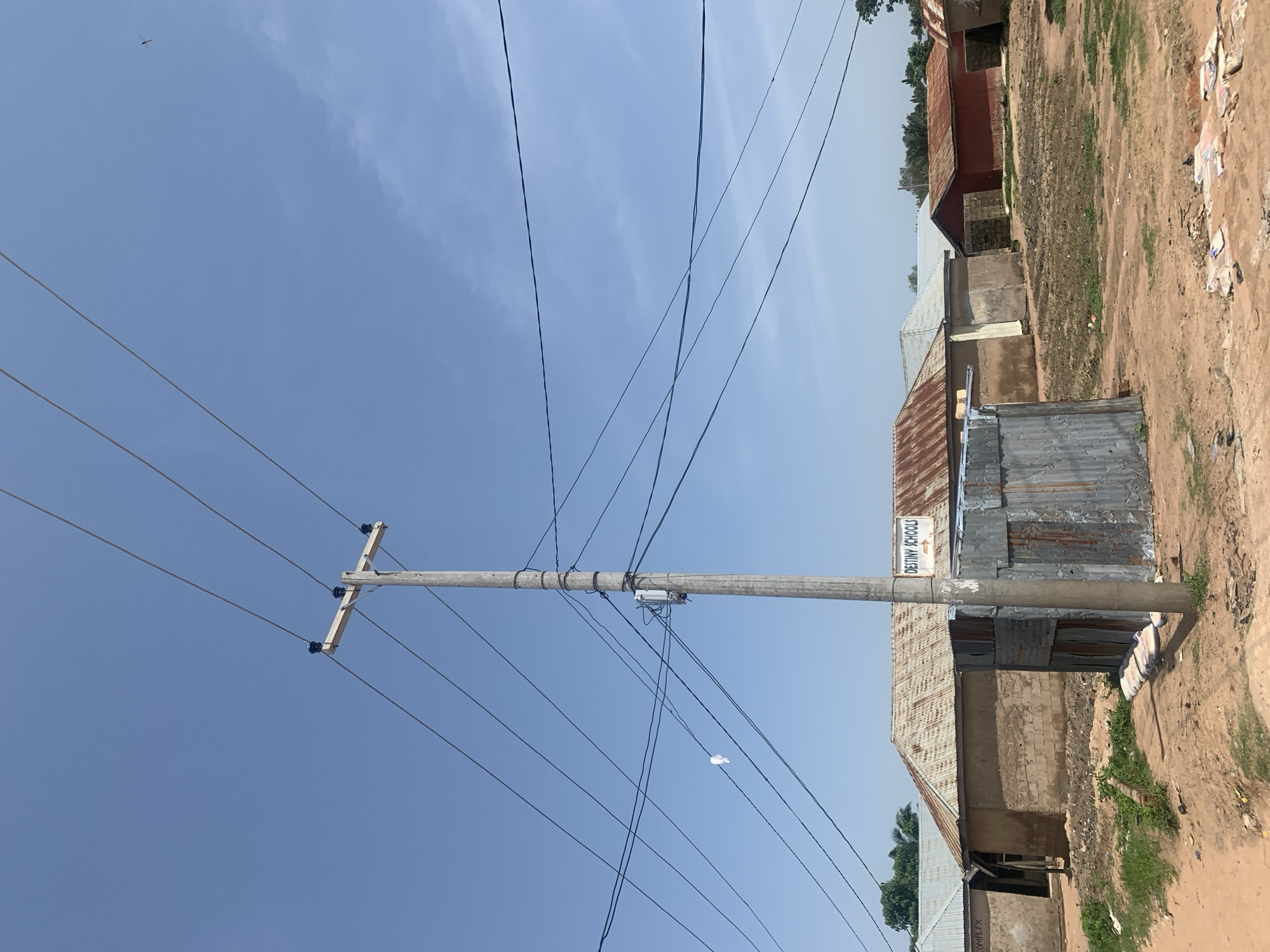 This image depicts a street scene in Jauro Yinu, Nigeria, under a clear blue sky. A prominent concrete utility pole stands in the foreground, supporting multiple overhead electrical wires that crisscross the frame. Attached to the pole is a small, white electrical box. To the right of the pole is a small, rustic structure constructed from corrugated metal. Behind these elements, several buildings are visible. One building, identified by a sign reading "DESTINY SCHOOLS" with an arrow, has a rusted corrugated metal roof and light-colored walls. Adjacent to it are other structures with similar roofing materials and varying wall constructions, some appearing to be made of mud brick. The ground is unpaved and appears to be composed of dirt and sand, with some scattered debris and patchy green vegetation. The overall impression is one of a semi-urban or rural setting, likely a residential or community area.