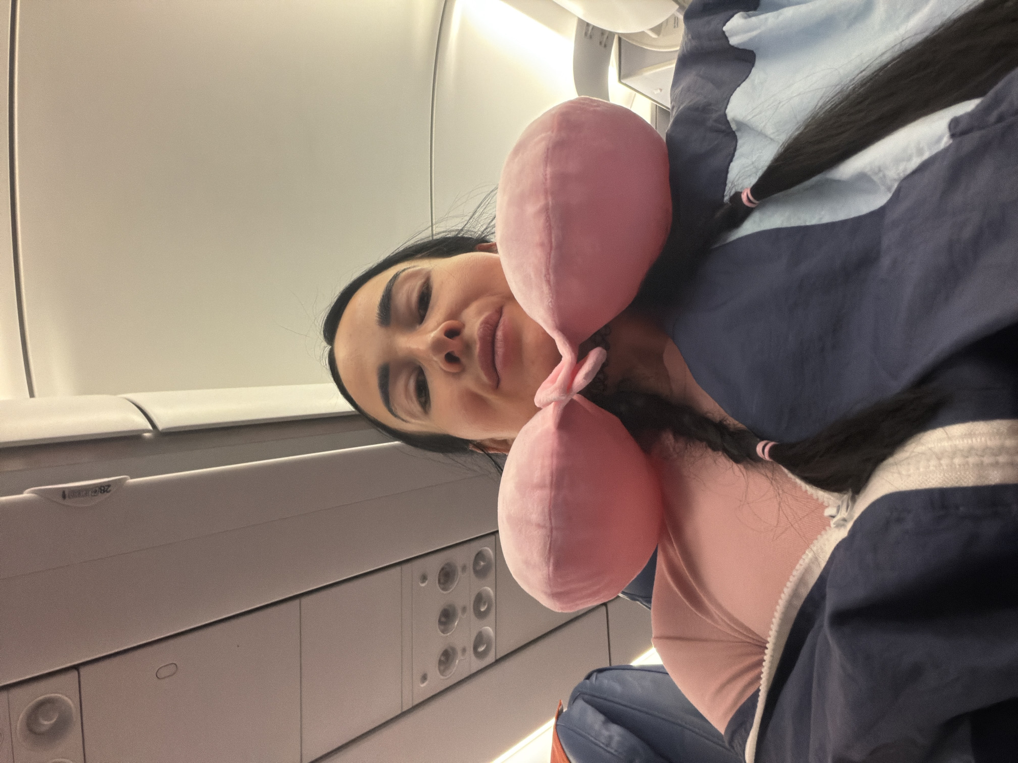 A woman is seated on an airplane, wearing a pink t-shirt and a dark blue and light blue jacket. She has a pink neck pillow around her neck and her dark hair is in two braids. The overhead compartment and lighting of the airplane cabin are visible in the background. The scene is likely in Saint Petersburg, Russia.