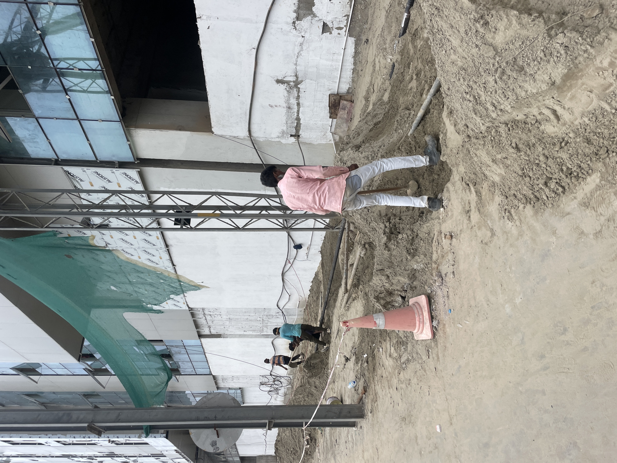 In New Delhi, India, at a construction site, three male laborers are engaged in digging. One man in a pink shirt and light-colored pants is using a shovel to dig into a mound of dirt. Two other men are working in a trench; one in a blue shirt and dark pants appears to be pulling a rope, while the other in a striped shirt is carrying something. The environment is dusty with piles of soil, construction materials, and exposed pipes. A construction building with glass windows and white panels is visible in the background, partially covered by green netting. A traffic cone is also present in the foreground.