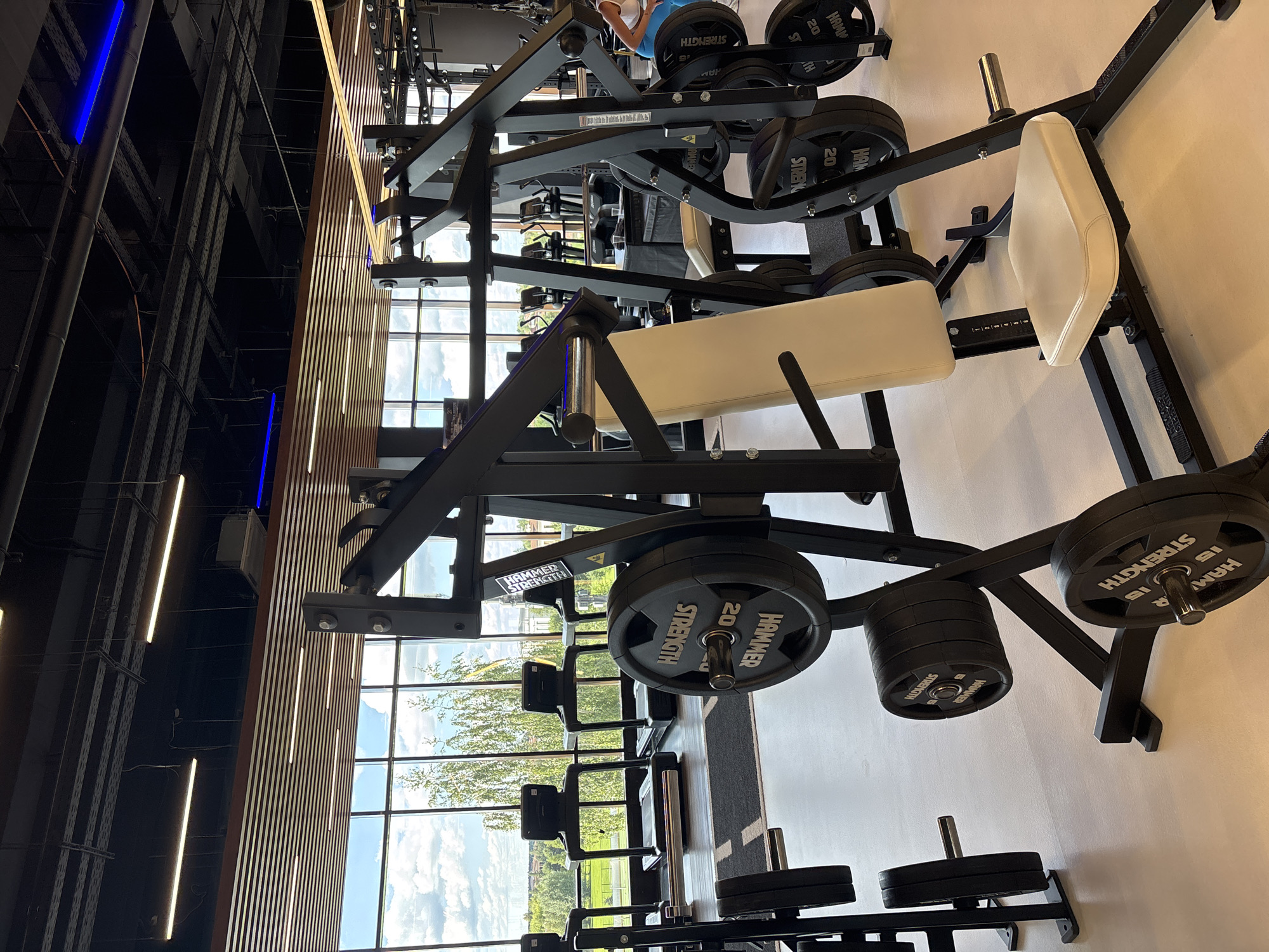 The image depicts the interior of a modern gym, likely located in Monolit, Russia. The space is well-lit with fluorescent and blue LED lighting fixtures visible on the ceiling, which is adorned with wooden slat paneling and exposed industrial piping. Large windows offer a view of a green outdoor landscape with trees and buildings under a cloudy sky.
The foreground is dominated by weightlifting equipment. A black Hammer Strength weight machine with a beige padded seat and backrest is prominently featured. Several black weight plates with "HAMMER STRENGTH" and "20" markings are loaded onto the machine's racks. Another weight plate rack is visible to the left, also with black weight plates. In the background, multiple treadmills are lined up facing the windows.
A person, partially visible on the right side of the image, is wearing a white top and blue leggings and appears to be using or interacting with the equipment. The floor is a light-colored, polished surface. The overall impression is one of a clean, well-equipped, and spacious fitness facility.