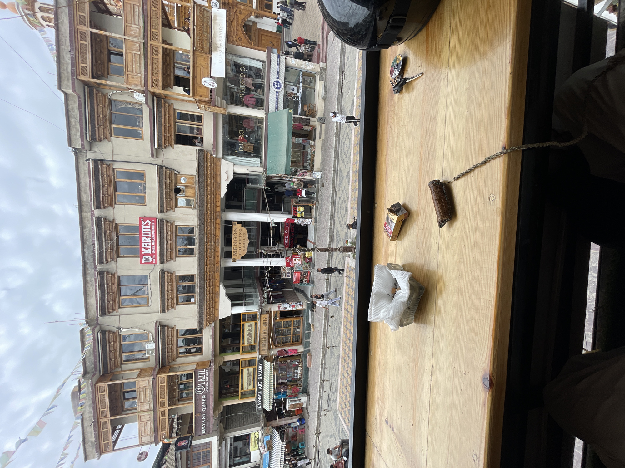 A high-angle, medium shot captures a street scene in Leh, India, viewed from a wooden-tabled interior. In the foreground, a wooden table displays an ashtray with a crumpled white paper lining containing cigarette butts, a partially smoked cigarette with a leather tip attached to a string, a pack of cigarettes, a set of keys, and the edge of a black motorcycle helmet. The background reveals a multi-story building with traditional Ladakhi architecture, featuring wooden balconies and intricately carved window frames. The building houses various shops and restaurants, including "Biryani Queen Lazil," "Kashmir Art Gallery," and "Men-gon Shopping Complex." Prayer flags are strung across the building. On the street below, pedestrians walk by, and several vehicles are parked. The sky is overcast.