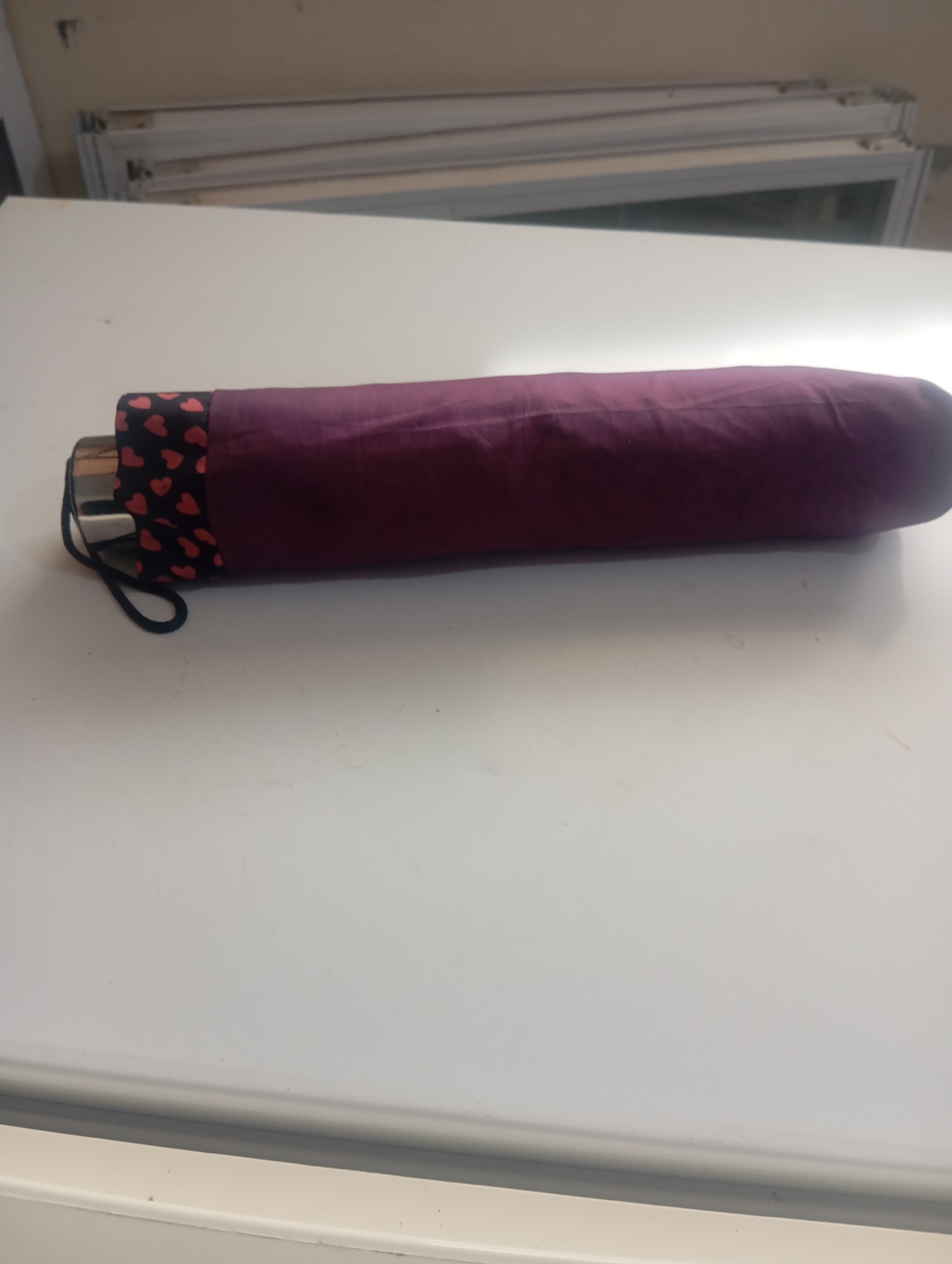 An umbrella is closed and lying horizontally on a white surface, possibly a table. The umbrella is covered in a deep red fabric with a pattern of small black hearts. The handle of the umbrella is visible on the left side and has a black strap attached. In the background, out of focus, are what appear to be window frames or shelving units. The image is taken from a slightly elevated perspective. The scene does not contain any identifiable features that would indicate a specific location within the city of Jos, Nigeria.