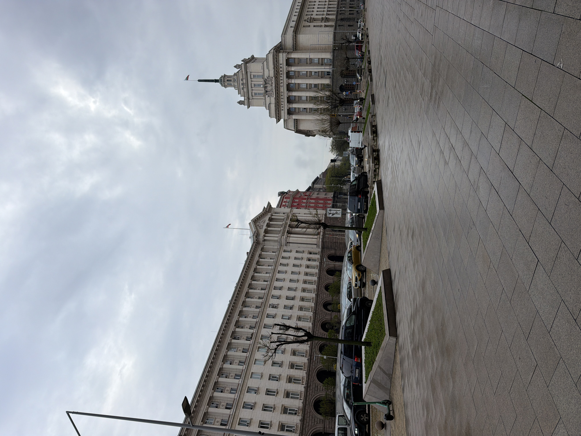 This media file depicts a wide street scene in Sofia, Bulgaria, under an overcast sky. The image captures two significant buildings, a paved pedestrian area, and parked vehicles.
The prominent building on the left (when correctly oriented) features a grand, imposing facade with a central tower and spire. Classical architectural elements such as columns and decorative moldings are visible. Clearly inscribed on its front is the text "НАРОДНО СЪБРАНИЕ," which translates to "National Assembly" or "Parliament" in Bulgarian. A small flag, likely the Bulgarian national flag, flies from a flagpole atop the tower.
To the right, a long, multi-story building with a uniform facade of numerous windows extends along the street. This building also displays classical architectural influences, with arched windows on its lower levels. Bare-branched trees are planted in green, raised planters along the street in front of it. Another small flag is visible on its roofline.
The foreground is dominated by a wide, gray-tiled pavement or sidewalk, which appears slightly damp, reflecting the dull light from the sky. A row of vehicles, including cars and vans, is parked along the street between the paved area and the buildings. Small patches of green grass are visible in the planters bordering the parking area.
The sky is entirely overcast, covered in gray clouds, suggesting a cloudy day with diffused light. No people are visible, and there is no discernible activity other than the static presence of buildings and parked cars. The scene conveys a quiet, institutional atmosphere in the capital city of Sofia.