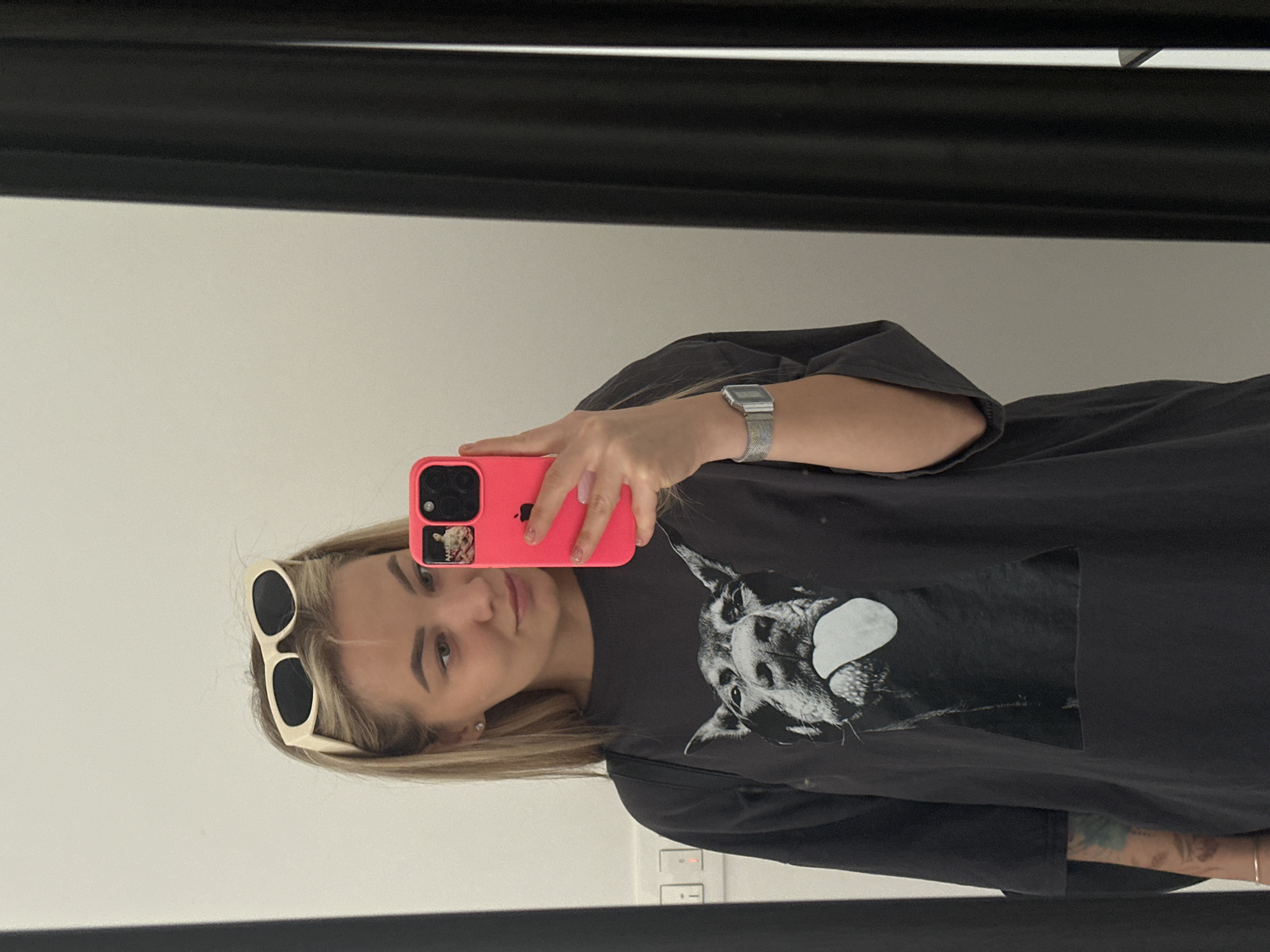 A young woman takes a selfie in a mirror. She holds a pink iPhone in her right hand. She wears sunglasses on her head, a gray t-shirt with a dog graphic, and a silver watch on her left wrist. The background is a white wall. The location is not discernible from the image.