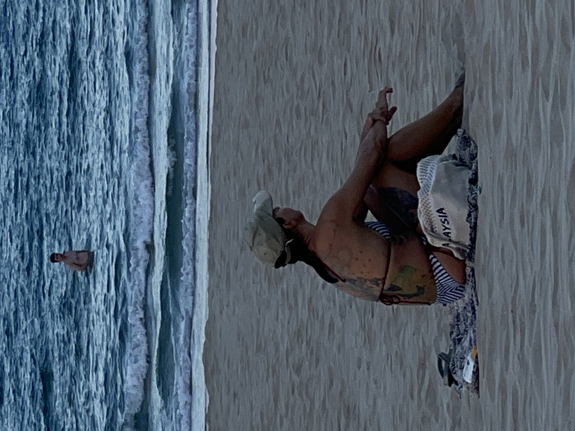 An adult male is standing in the shallow ocean water on a sandy beach. In the foreground, a woman with tattoos on her back is sitting on the sand facing away from the camera. She is wearing a bikini and a baseball cap. Next to her is a beach bag with the word "MALAYSIA" printed on it, a pair of flip-flops, and a towel. The ocean has gentle waves. The overall scene appears to be at a beach in Da Nang, Vietnam.