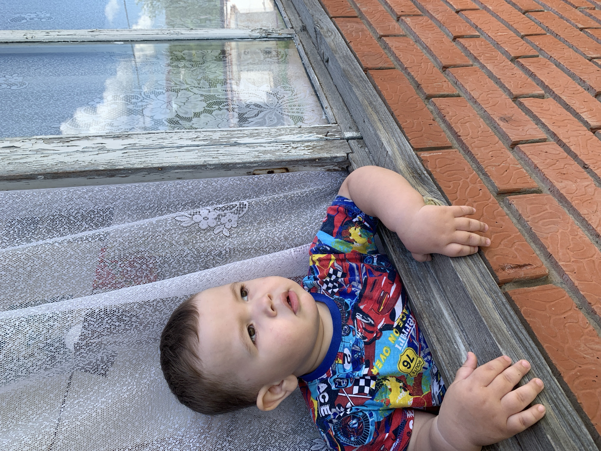 A young child is positioned in a window frame. The frame is bordered by a brick wall on one side and a window with peeling paint and lace curtains on the other. The child is wearing a colorful shirt with cartoon car designs. The reflection of trees and sky can be seen in the window panes. The scene takes place outdoors in Фоки, Russia.
