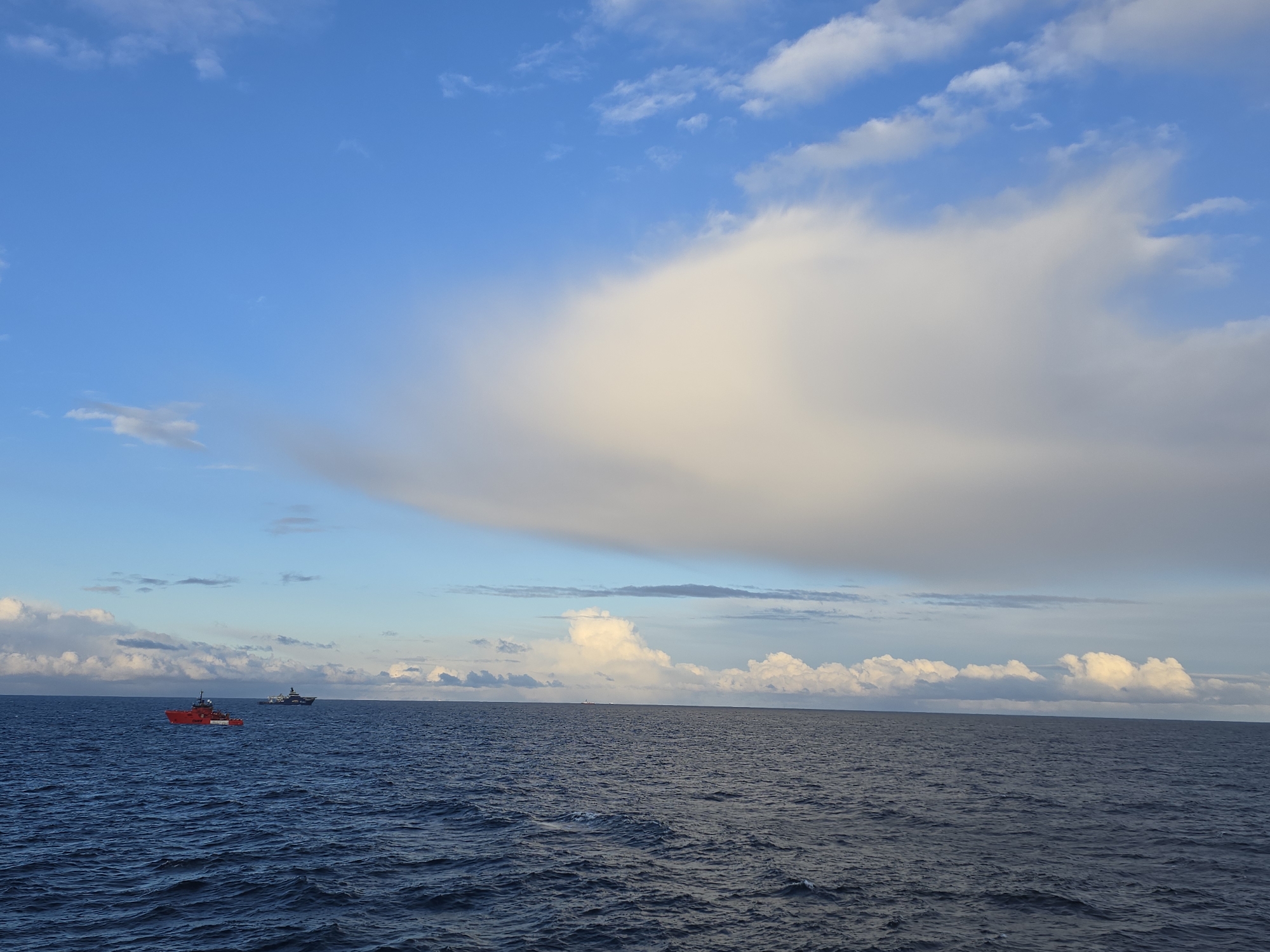 The image shows a seascape under a blue sky with scattered clouds. The sea is dark blue and slightly choppy. There are two ships visible in the distance. The closer one is painted red and white, while the other is a darker color. The location is Unknown, Unknown. There are no people visible.