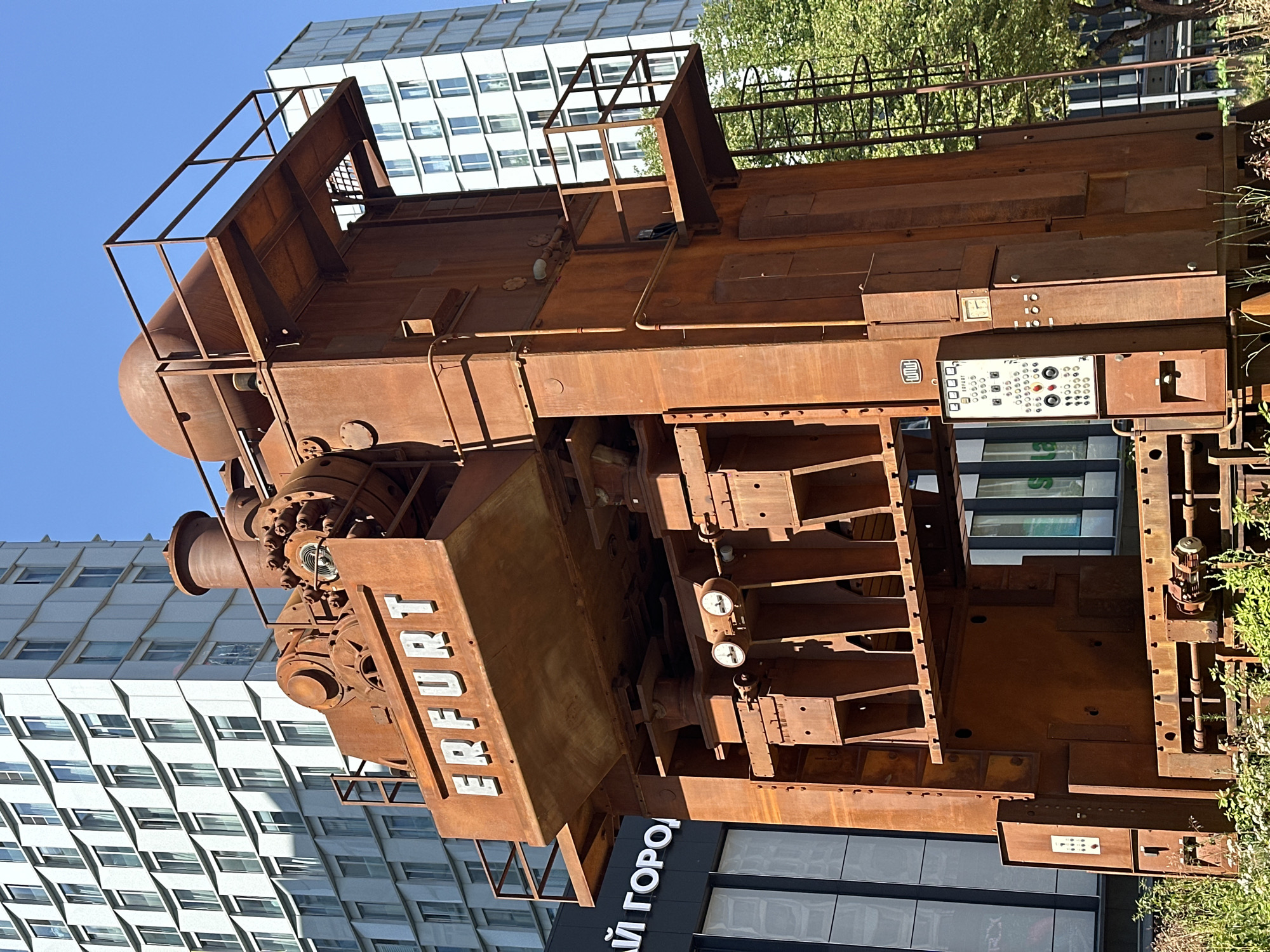 The image shows a large, rusty industrial press with the name "ERFURT" prominently displayed on its upper section. It is situated outdoors in an urban environment, with modern, white buildings in the background. The press is made of weathered metal and features various mechanical components, including pipes, platforms, and a control panel with buttons and a clock. Green foliage is visible at the base of the structure and to the right. To the left of the press, a building with dark facade and white lettering that appears to say "ЛУЙ ГОРОД" is partially visible. This industrial press is located in Moscow, Russia, specifically near the former "Krasny Proletariy" (Red Proletarian) factory, which produced machine tools. The sculpture is a remnant of the factory's industrial past, now repurposed as a public art installation.