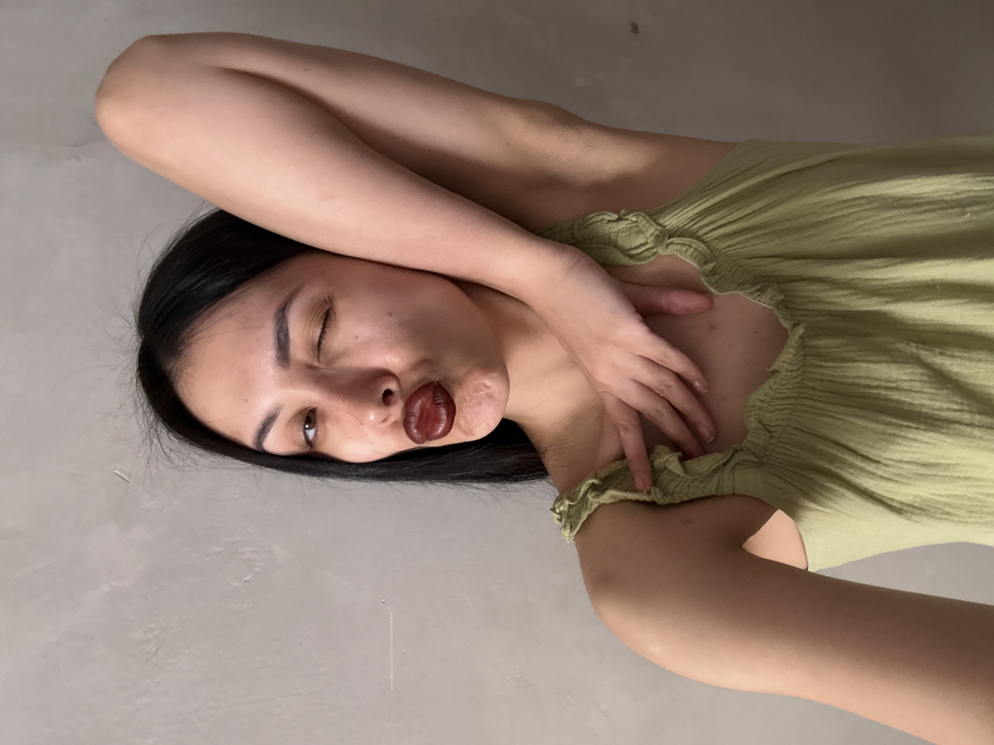 A woman with dark hair lies on her back against a gray wall. She is wearing a light green, textured, ruffled top. Her arms are crossed above her head, with one hand near her face and the other resting on her chest. She is winking one eye and puckering her lips. There is no indication that the photo was taken in Votkinsk, Russia.