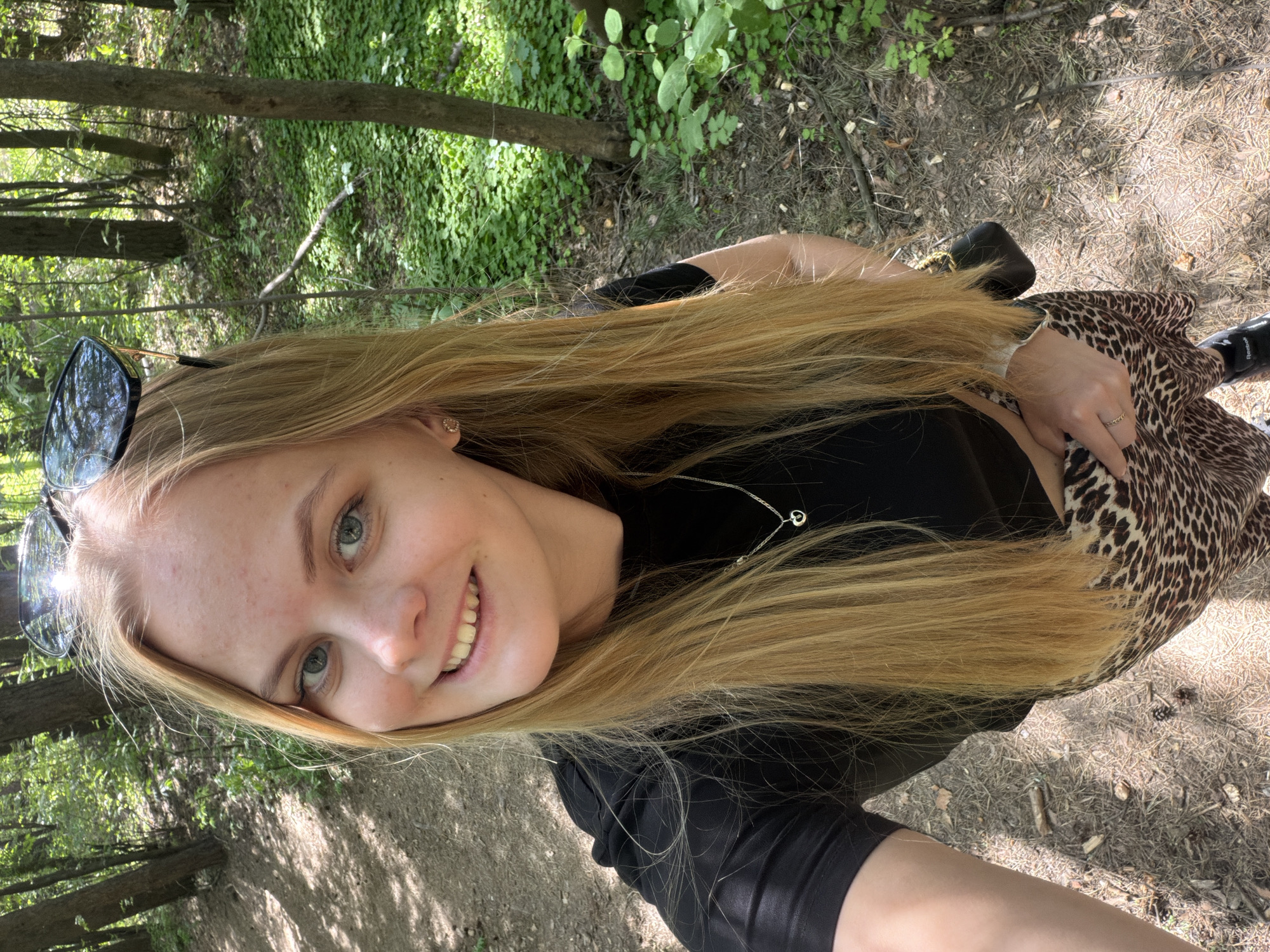 A young woman with long blonde hair is posing for a photograph in a wooded area. She is wearing sunglasses on her head, a black shirt, and a leopard-print skirt. She is smiling and looking at the camera. The background shows trees and ground covered with leaves and twigs. The location is possibly Izhevsk, Russia.