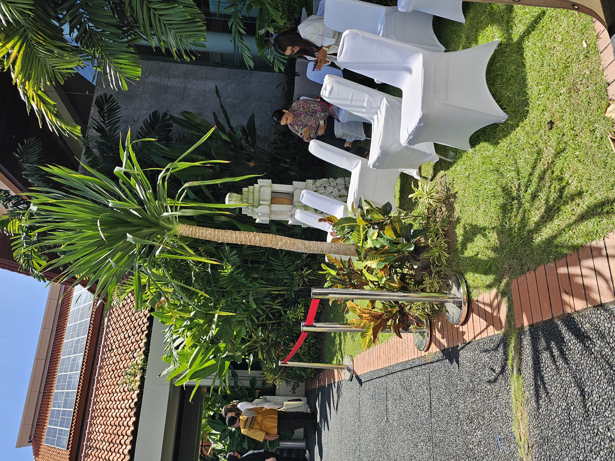 The image depicts an outdoor event setting in Kuta Selatan, Indonesia. On the left is a building with red tiled roofing and a window pane that reflects the blue sky. A tall tree with long green leaves stands adjacent to the building, extending towards the center.
Chairs covered in white fabric are arranged on the right on a patch of green grass. There are two people in the image sitting on the chairs. One is wearing a white top, and the other is wearing a floral shirt.
In the center, a white stone structure is set as a focal point among dense tropical foliage. A red velvet rope is held up by two silver stanchions. The pathway is made of small dark gray pebbles and leads to a brick border. The lighting suggests a sunny day, with strong shadows cast by the tree and stanchions. Another group of people is visible in the distance, presumably attending the event.