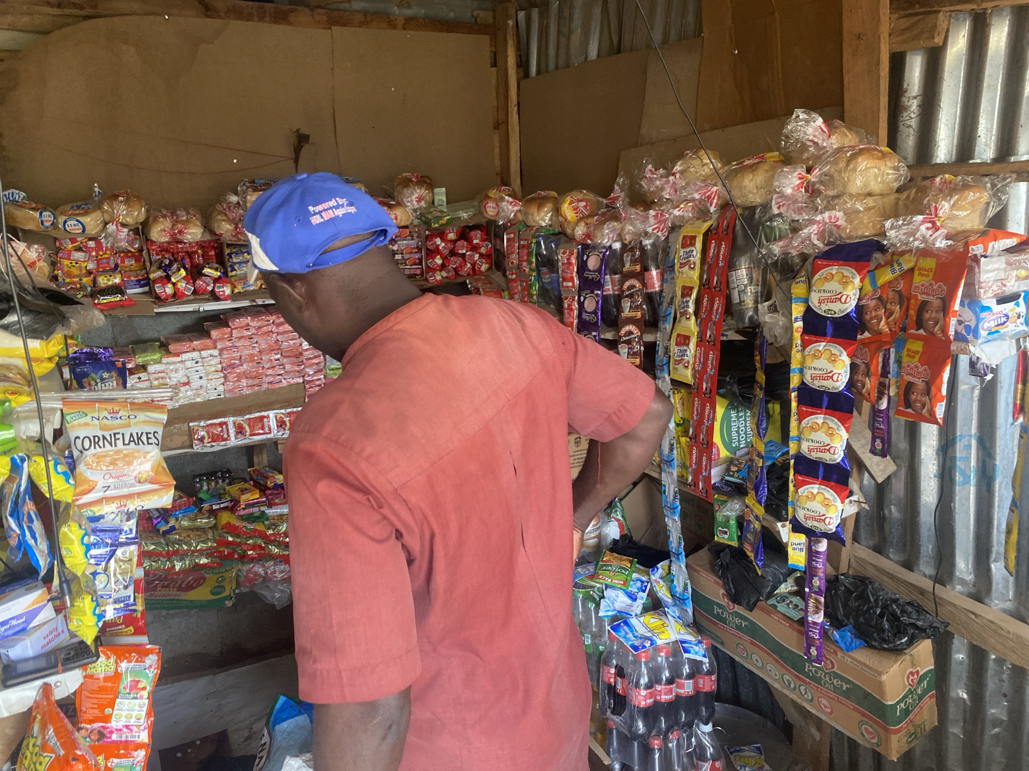 The image depicts the interior of a small, bustling market stall or kiosk, likely in Abuja, Nigeria. A person, seen from behind, is the main subject, wearing a reddish-orange short-sleeved shirt and a blue baseball cap. Text on the cap reads: "Powered By: HON. MAM Agailaapa". The person appears to be browsing or organizing the numerous items on display.
The stall's structure is simple, featuring walls made from corrugated metal sheets, wooden planks, and brown board. The space is tightly packed with a wide variety of consumer goods. Products are arranged on shelves, hung from strings, and stacked on the floor.
Visible merchandise includes several loaves of bread wrapped in clear plastic bags, "NASCO CORNFLAKES" cereal, and multiple packets of "Danish COOKIES." Other identifiable food items are "SHACK Pack" snacks, "PARACO Milk," "SUPREME NOODLES," "MILO," and "Three Crowns" sachets. Non-food items include "closeup" toothpaste and "ger Head safety matches." Bottles of soda, likely Coca-Cola, are stacked in plastic carriers near the bottom right, next to a cardboard box labeled "Power Oil." "Pan Custard" and "Macaroni" are also visible. The bright illumination suggests it is daytime.
