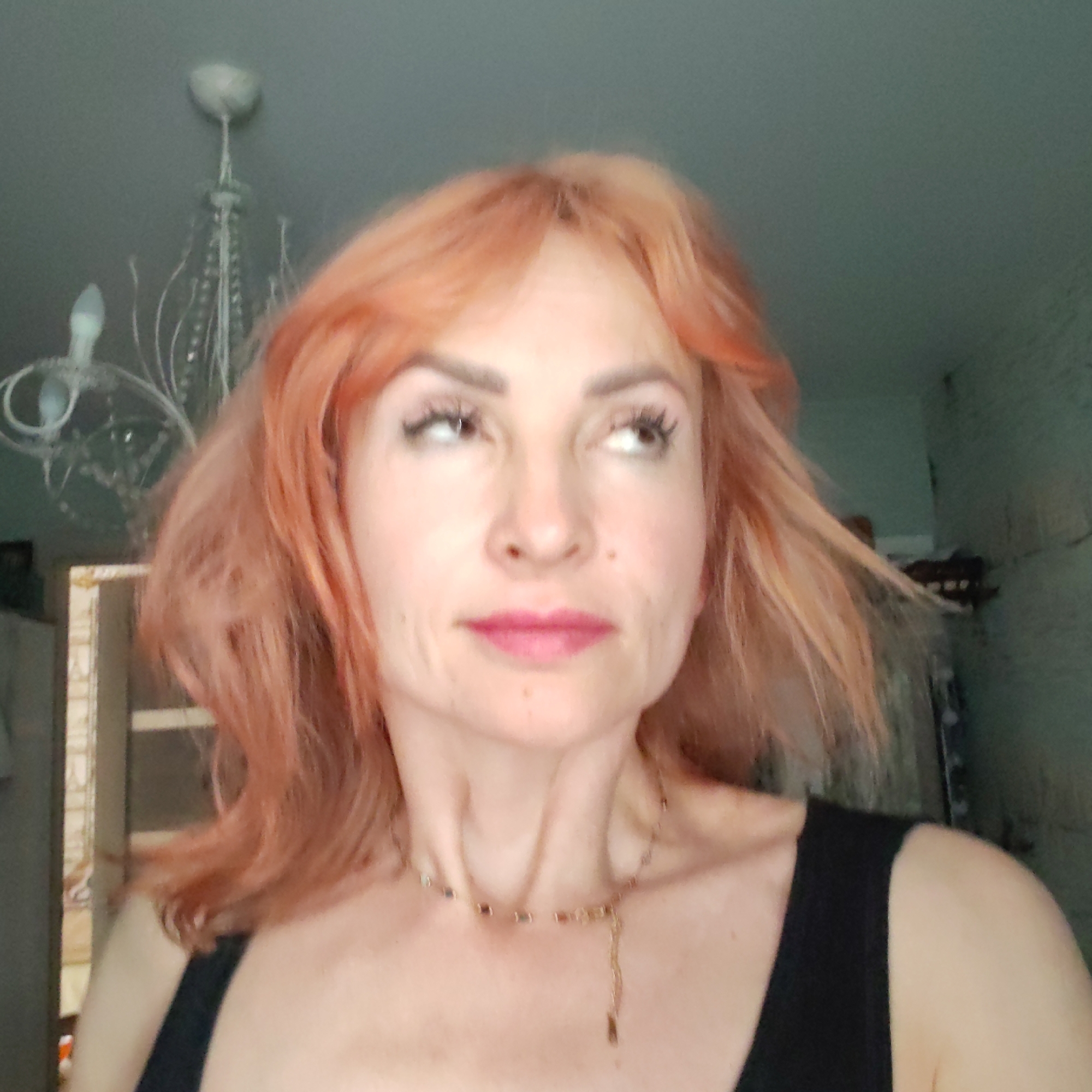 The image presents a close-up portrait of a woman with light reddish-orange, shoulder-length hair styled with bangs. She has fair skin, dark eyebrows, prominent eyelashes, and her lips are adorned with pink lipstick. Her gaze is directed upwards and to her right, giving her a pensive or thoughtful expression. She is wearing a black tank top and a delicate, gold-toned necklace featuring small beads or charms around her neck.
The setting is indoors, with elements of a room visible in the background. In the upper left corner, a white or light-colored chandelier with multiple arms and crystalline adornments hangs from the ceiling. A light bulb is visible on one of its arms. To the left, a blurred portion of what appears to be a doorframe or window frame suggests another part of the room. The right side of the background features a textured wall, possibly painted in a muted teal or greenish-blue hue, with some indistinct darker objects or furniture partially visible against it. No text is visible in the image.
