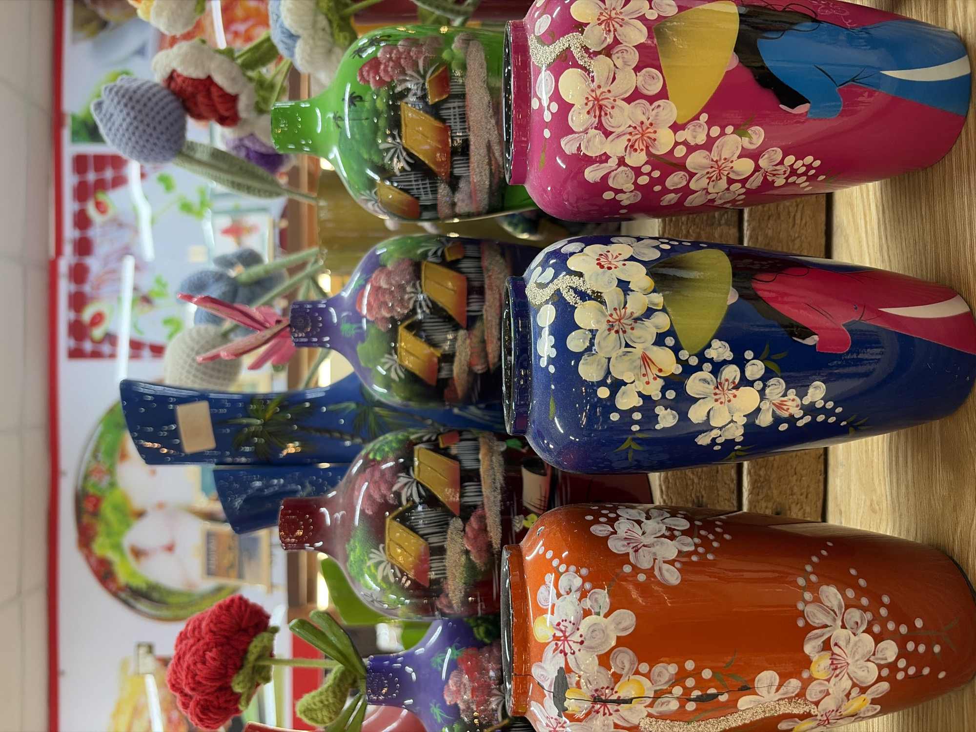 The image shows a close-up, eye-level shot of an assortment of decorative vases displayed on a wooden surface. The vases are arranged in two rows. The front row features three vases: an orange vase on the left adorned with white flowers and a black umbrella, a blue vase in the center with white flowers and a depiction of a woman in traditional Vietnamese attire, and a pink vase on the right with similar white floral decorations and a representation of a woman in traditional dress. The second row, positioned behind the front row, displays larger vases. From left to right, there is a purple vase, a dark red vase with a landscape painting of houses and palm trees, a blue vase with a palm tree motif, and a green vase featuring a similar landscape painting. In the background, more vases and decorative items are visible, including knitted flowers in various colors. The overall scene suggests a retail environment, possibly a souvenir shop or a craft market in Da Nang, Vietnam, known for its vibrant art and craft scene. The lighting is bright, illuminating the glossy finishes of the vases. There are no people clearly visible in the image.