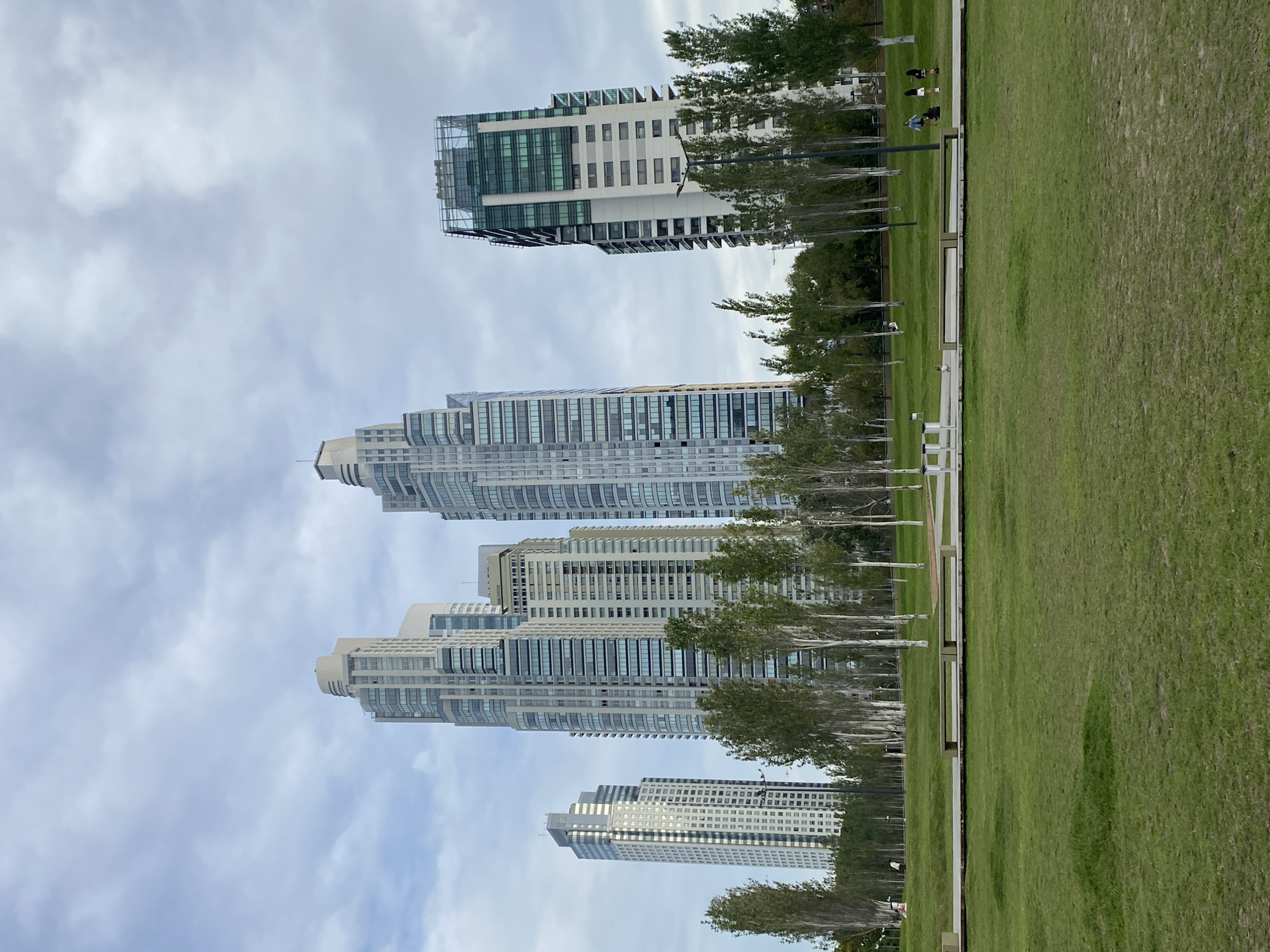 The image shows a park in the Puerto Madero neighborhood of Buenos Aires, Argentina. Four modern skyscrapers of similar design line the left side of the image. In between the buildings are several thin trees. To the right, a large expanse of green lawn is visible, dotted with benches and pathways. In the distance, several people are sitting on the grass, engaged in conversation or relaxation. The sky is overcast with a mix of clouds and patches of blue.