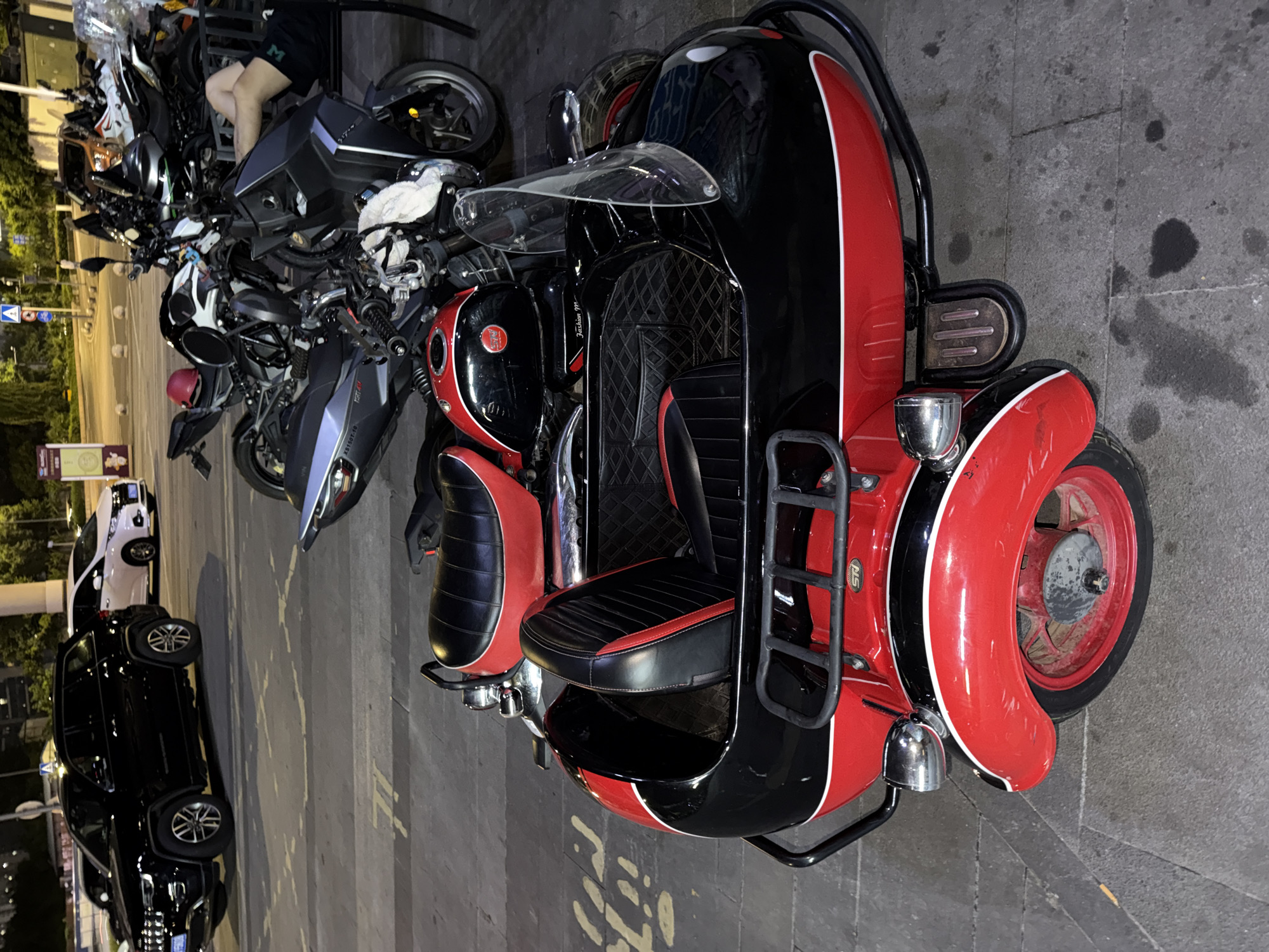 A black and red motorcycle with a sidecar is parked on a paved street in the city of Chongqing High-tech Zone, China. The motorcycle has a black fuel tank with red accents and a red seat. The sidecar is also black and red, with a black upholstered seat. A person is partially visible in the sidecar. In the background, other motorcycles and a black SUV are parked. The lighting suggests it is evening or nighttime.