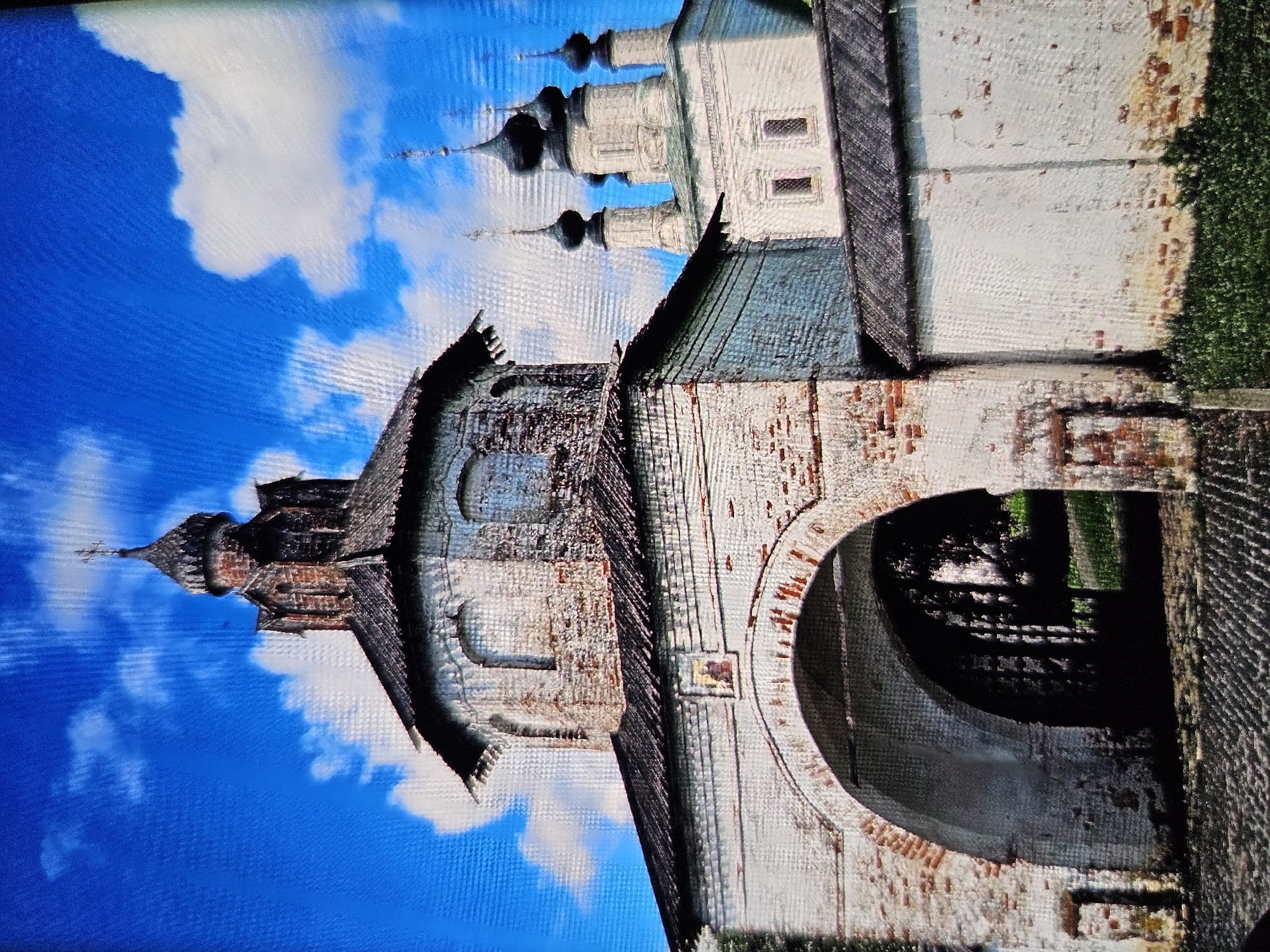 The image shows a view of the Golden Gate in Vladimir, Russia, on a bright, sunny day with a blue sky and scattered white clouds. The Golden Gate is a historical monument, a triumphal arch built in the 12th century. It is constructed of white stone and brick, with a pitched roof and a small tower topped with a cross. The archway is wide and dark, with a glimpse of green foliage visible through it. To the right of the Golden Gate, a white church with several dark domes is visible, also adorned with crosses. The foreground consists of a cobblestone path. The overall impression is one of ancient architecture set against a clear, vibrant sky.