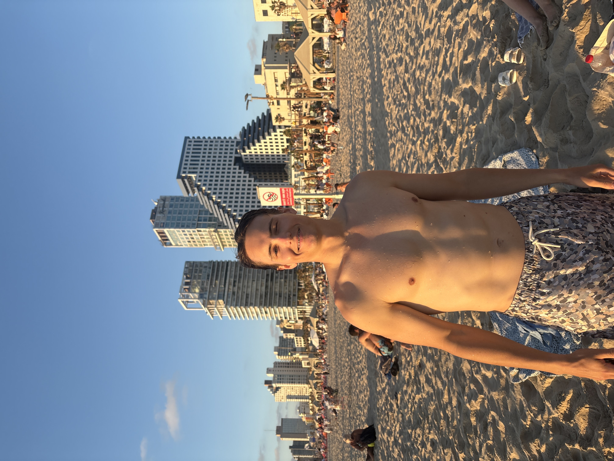 A young man, appearing to be in his late teens or early twenties, stands on a sandy beach in Tel Aviv, Israel, facing the camera. He is shirtless, with water droplets visible on his skin, and is wearing patterned swimming shorts. The background features a clear blue sky with a few wispy clouds. Behind the man, a line of modern high-rise buildings, characteristic of the Tel Aviv skyline, stands against the horizon. The beach is populated with numerous people, some sitting on towels or chairs, others walking, and several covered beach structures are visible. The sunlight suggests it is late afternoon or early evening. The overall scene depicts a casual, public gathering on a beach in an urban environment.