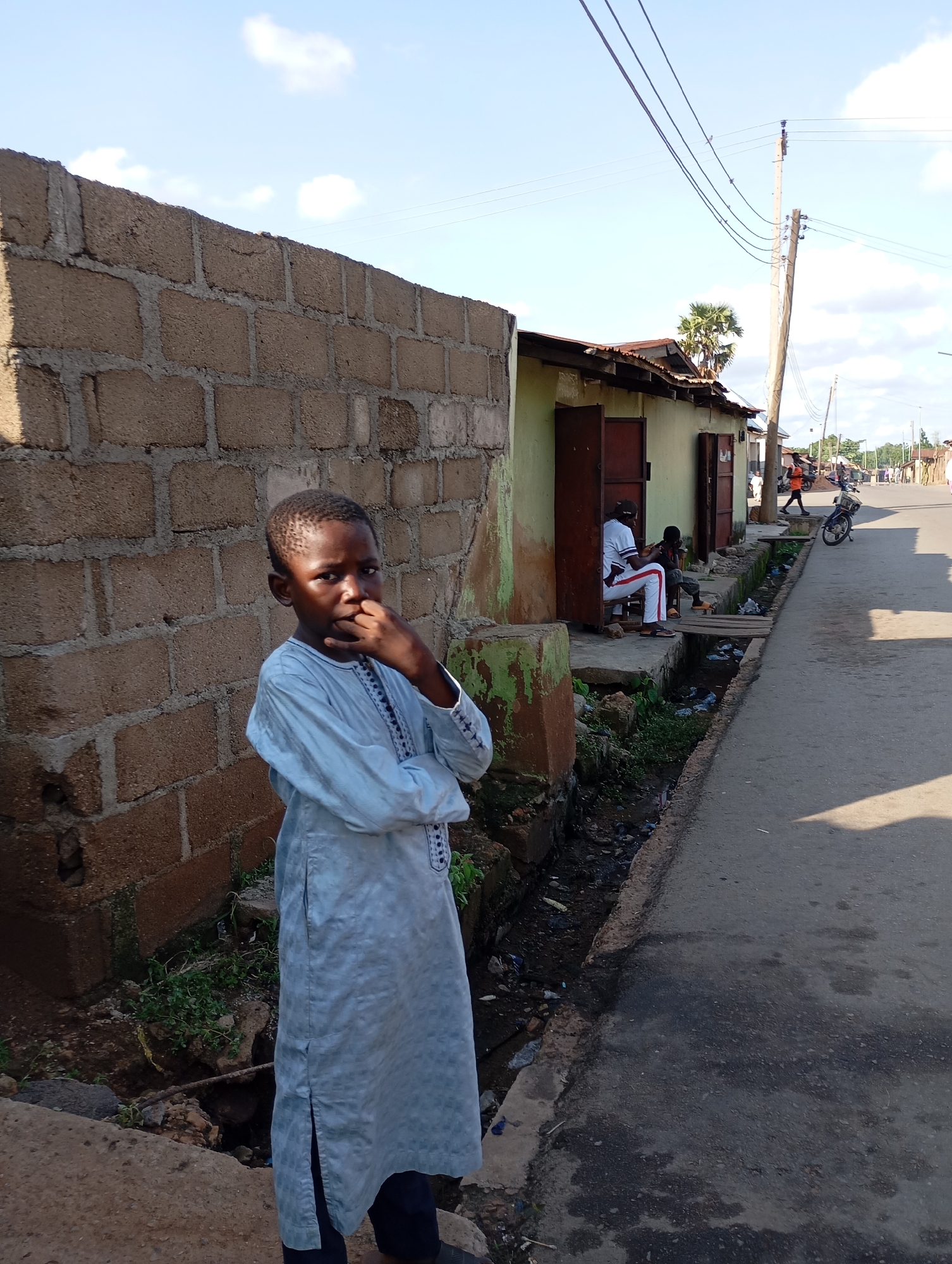 In Shendam, Nigeria, a young boy stands in front of a rough-hewn brick wall. He is wearing a light blue traditional garment with embroidered details around the neckline and cuffs. His arms are crossed, and his right hand is raised to his mouth, as if in thought or contemplation. The boy's gaze is directed towards the camera. To his right, a street runs parallel to the scene, with a shallow, debris-filled drainage channel running alongside it. In the background, a light green building with open doorways is visible, and two individuals are seated outside it. Further down the street, other people and a motorcycle can be seen, suggesting a bustling, everyday urban environment. The sky above is a clear blue with a few scattered white clouds, and utility wires stretch across the frame, supported by wooden poles. The overall impression is a snapshot of daily life in a Nigerian city.