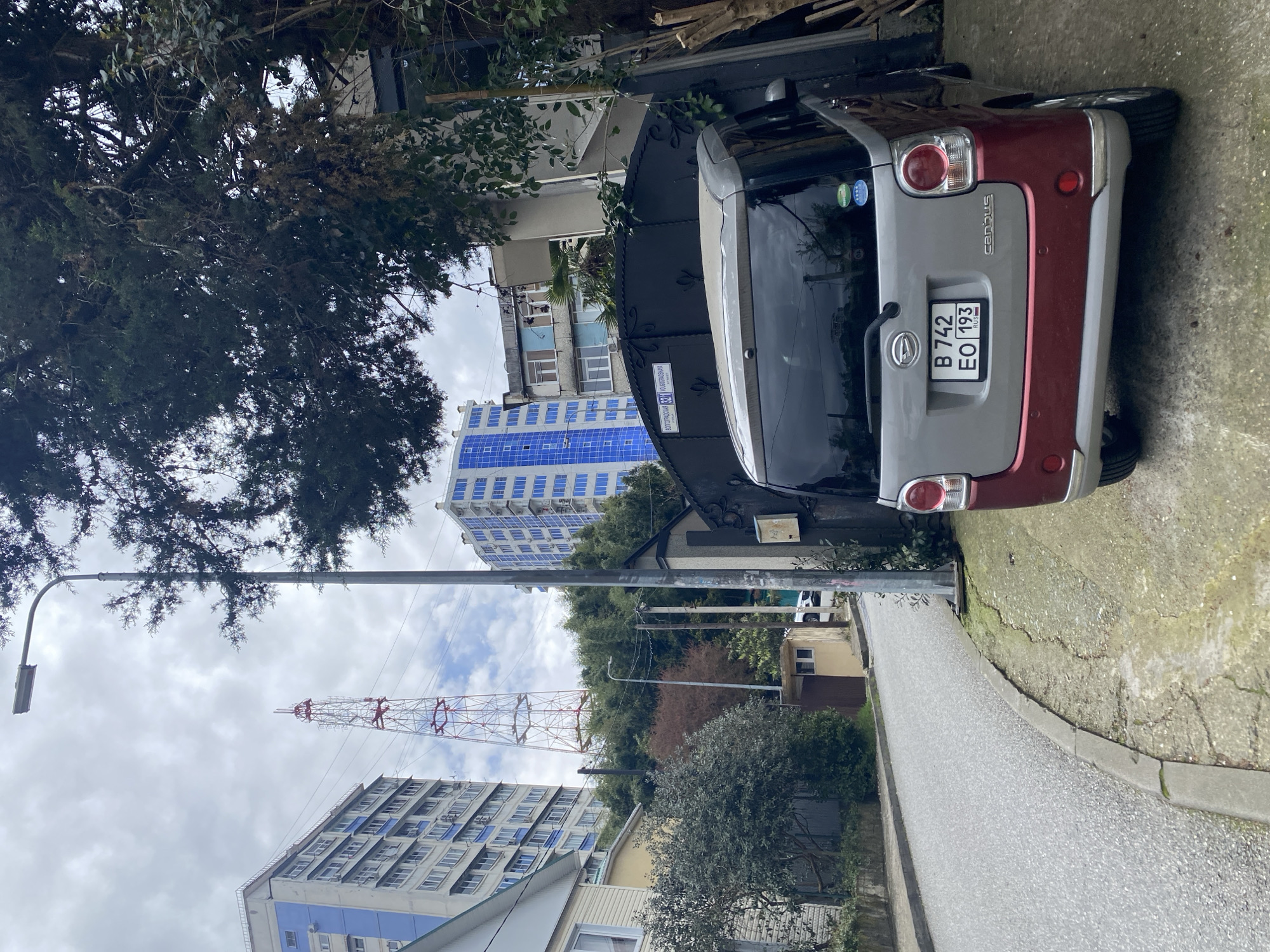 The image shows a residential street in Sochi, Russia, with a two-tone silver and red Daihatsu Canbus parked on the side. The car's license plate reads "B 742 EO 193" and indicates a Russian registration. In the background, several multi-story residential buildings are visible, one featuring distinctive blue-framed windows. A lattice communication tower rises near these buildings. A streetlight stands along the street, and a tree overhangs the scene. The street surface is concrete or asphalt, with a curb. The sky is overcast.