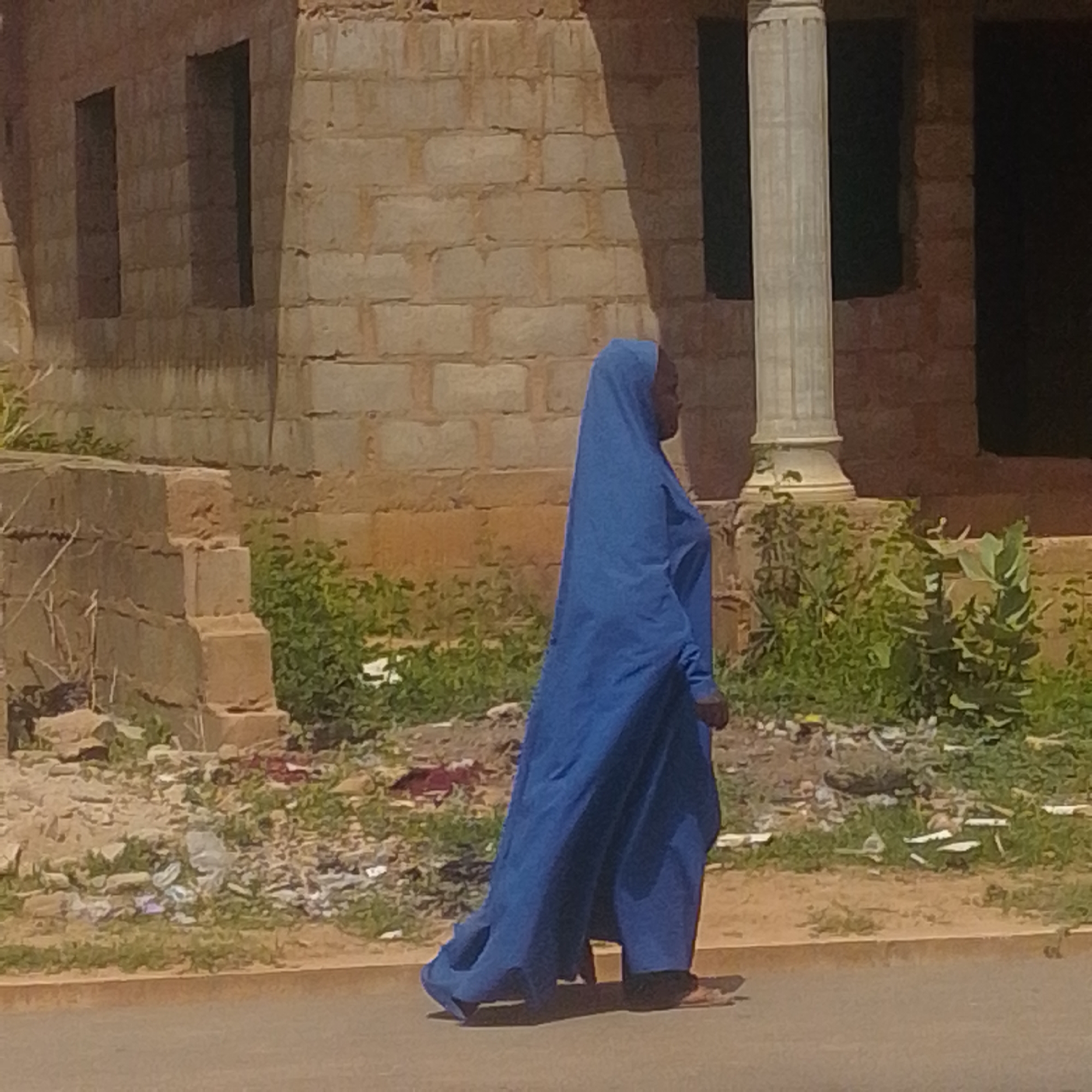 A person wearing a flowing blue garment and headscarf walks along a paved street in Gombe, Nigeria. The person is seen from the side, with their face partially obscured by the headscarf. The background features a brick building with two dark openings, possibly windows or doorways, and a large, fluted column to the right. In front of the building, there is dense green vegetation and scattered debris. The street is a dull gray, with a visible curb. The overall scene suggests an outdoor environment in an urban or semi-urban setting within Gombe.