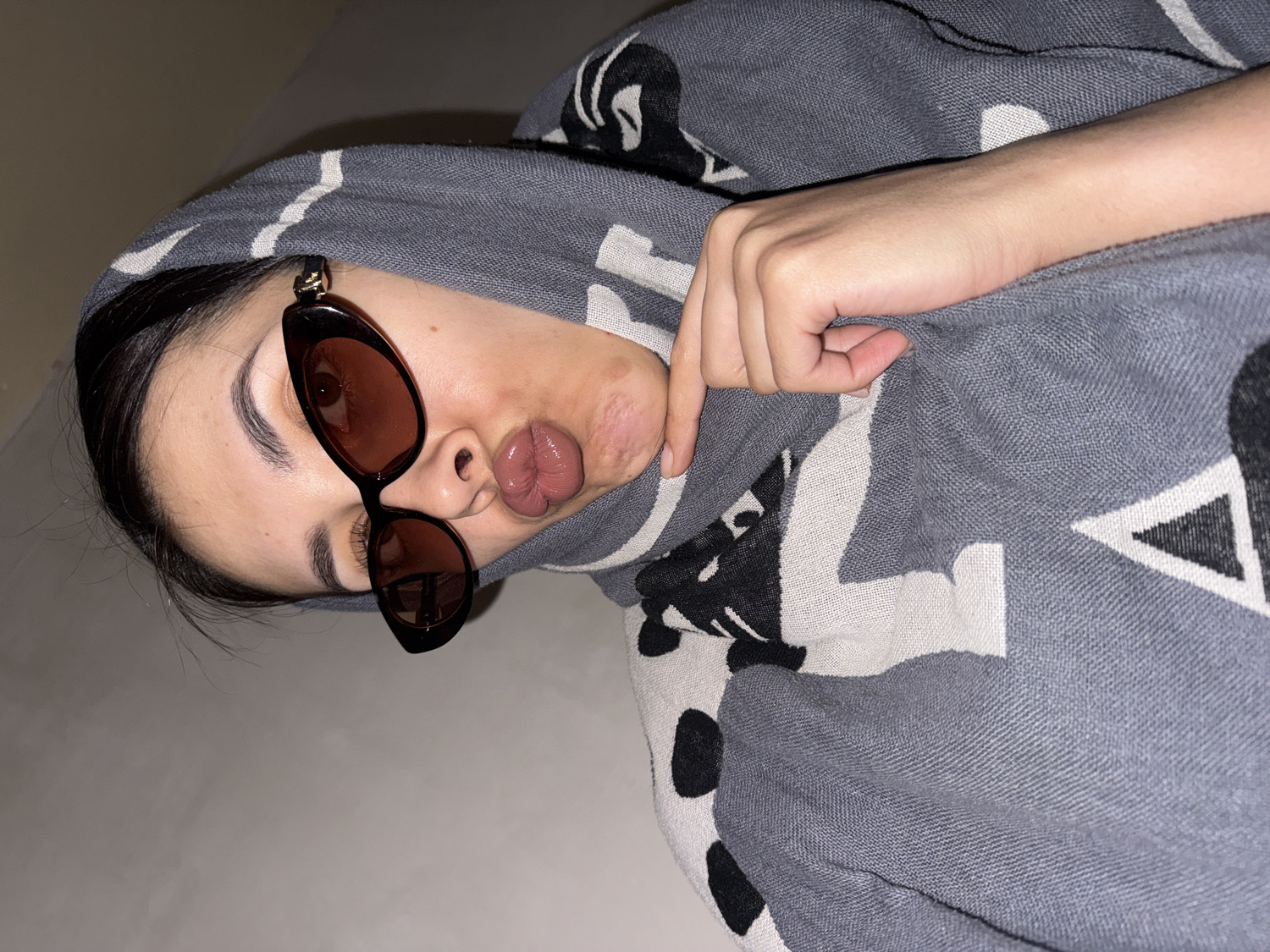 A headshot of a person, likely female, is presented, rotated 90 degrees counter-clockwise, with their head positioned on the left side of the frame. The individual wears dark brown, cat-eye style sunglasses with amber-tinted lenses, through which one eye is visible. Their lips are prominently puckered in a "kiss" or "duck face," displaying natural texture.
The person is cozily wrapped in a grey blanket or scarf that features a distinct, abstract pattern of black and white shapes, including triangles and other geometric and organic forms. A bare hand with visible fingers and a closed fist is resting on the blanket in the mid-right portion of the image.
The background consists of a plain, light-colored surface, suggesting an indoor setting, possibly a home. The lighting is soft and appears to be artificial, indicative of evening or night-time conditions, or simply a dim indoor environment. The overall mood conveyed is relaxed and playful. There is no visible text in the image. The location context provided is Votkinsk, Russia.