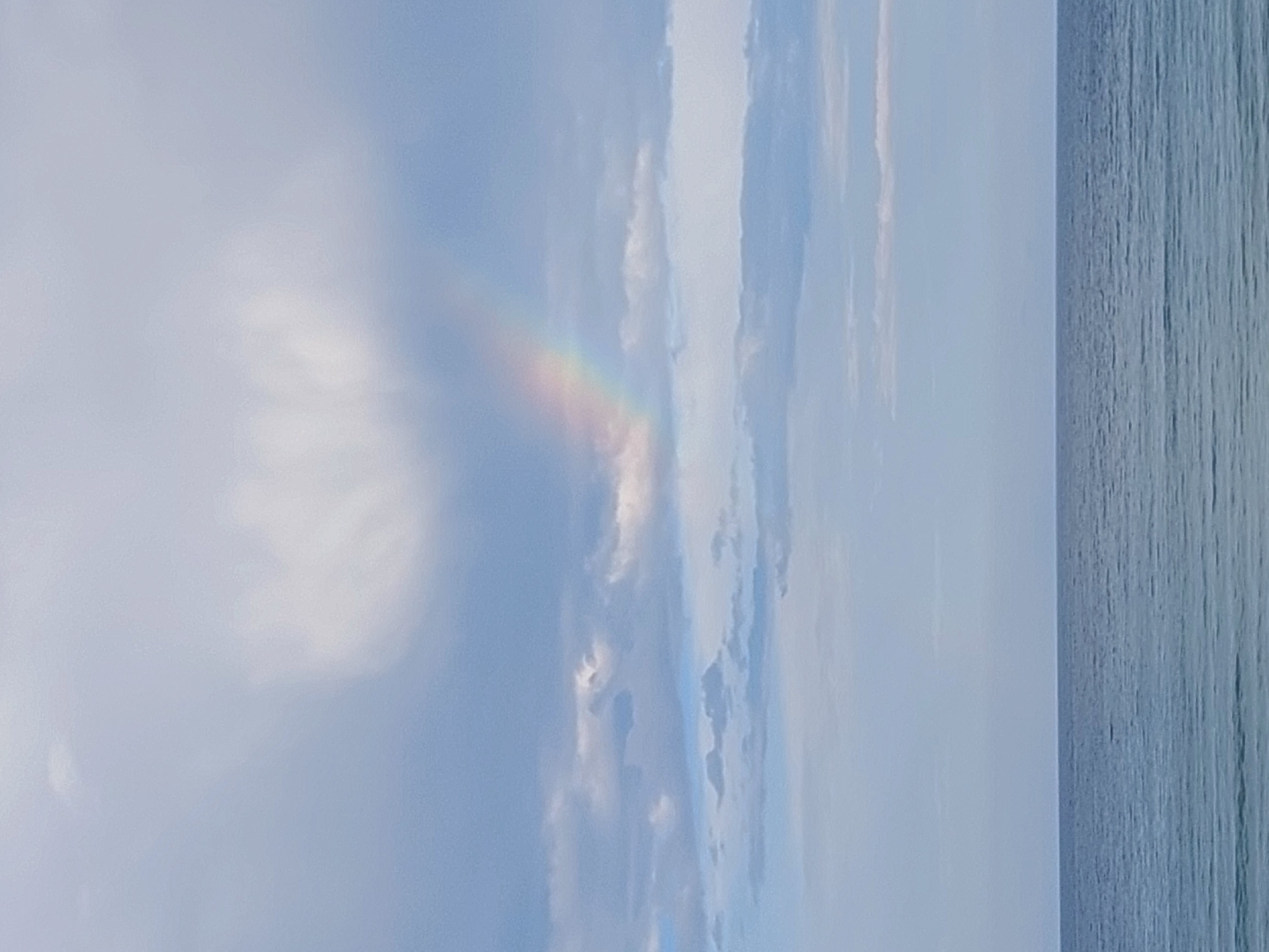 The image is a vertical, low-angle shot of a sky with a rainbow and the sea. The sky is mostly light blue with scattered white clouds. A faint rainbow arc is visible in the upper left quadrant of the image, with colors ranging from red to violet. The sea occupies the right side of the frame, appearing as a textured, deep blue expanse with subtle lines suggesting waves. The lighting indicates it's daytime, likely either early morning or late afternoon given the soft light and the presence of a rainbow, which typically forms when the sun is low in the sky and shining on water droplets. The overall mood is serene and natural. No people, objects, or text are visible. The location context is Sozopol, Bulgaria.