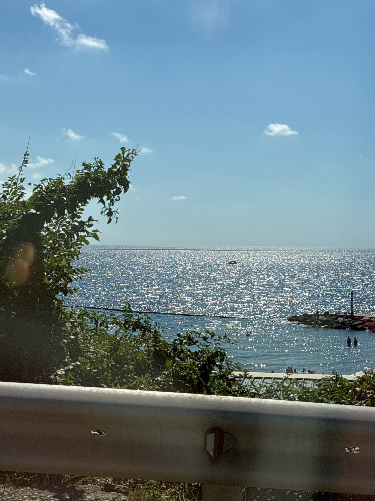 This image depicts a sunny day at a beach in Lermontovo, Russia. The scene is dominated by the bright, sparkling sea under a clear blue sky with a few scattered white clouds. In the foreground, lush green foliage partially obscures the view of the water and a portion of a silver guardrail. The sea is calm, with gentle waves and a distant horizon line. Several people are visible in the water and near a rocky jetty on the right side of the frame, some appearing to be swimming or wading. A small boat is visible on the water in the middle distance. The overall impression is one of a typical summer day at the seaside.