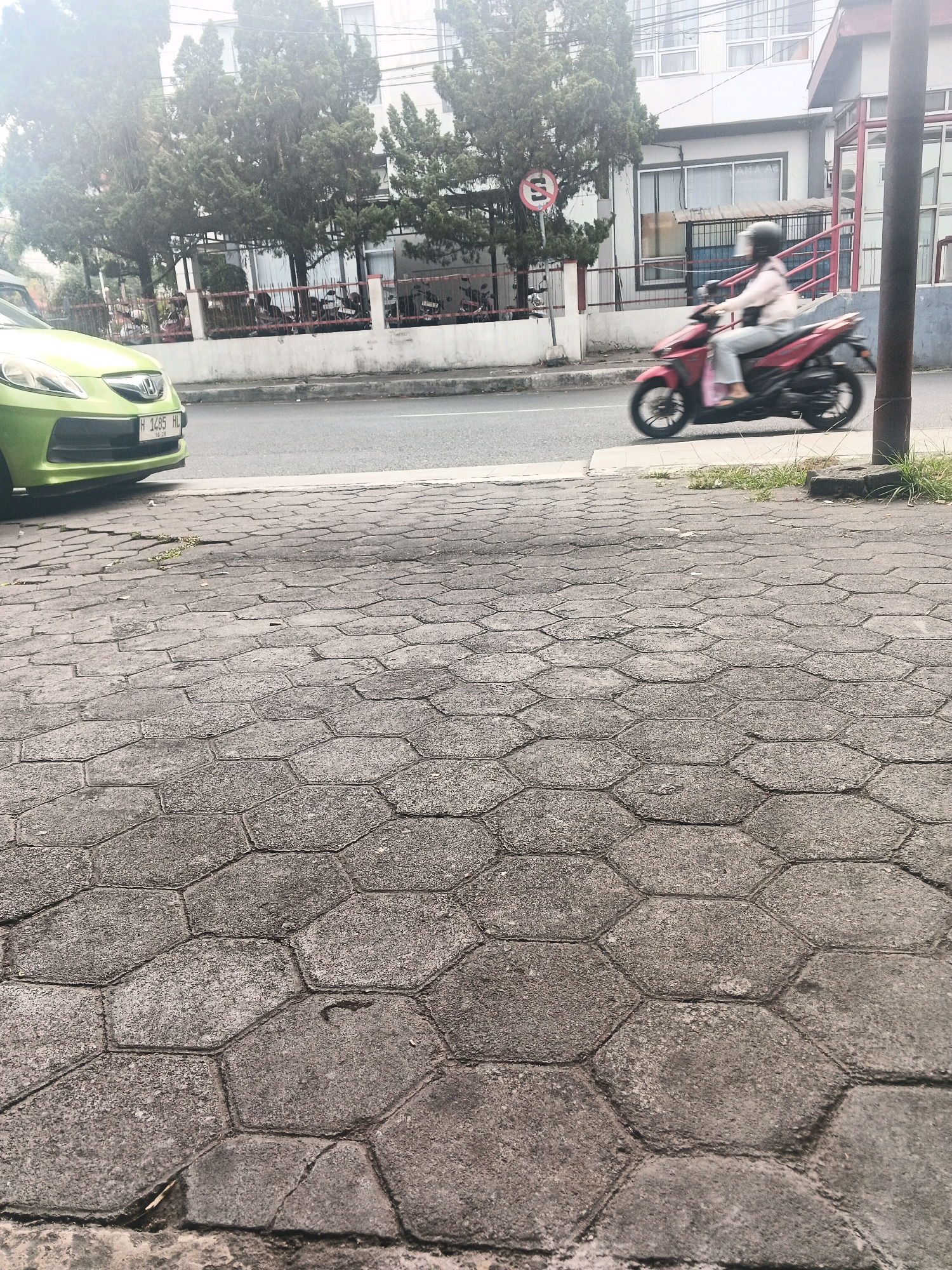 The image depicts a street scene in Ungaran, Indonesia. In the foreground, a grey hexagonal paver sidewalk fills the lower portion of the frame. The pavers are somewhat worn and cracked, with some debris scattered on them. To the left, the front of a bright green car is visible, partially obscuring the sidewalk. Its license plate reads "H 1485 HL".
Beyond the car and sidewalk, a paved road stretches across the middle ground. A person on a red motorcycle is moving from right to left across the road. The rider is wearing a helmet and a light-colored jacket over dark pants, and the motorcycle is blurred due to motion.
In the background, there is a building with a white facade and several windows, possibly an office or residential building. Trees with dense foliage line the area between the road and the building, and a red and white fence runs along the property line. A circular traffic sign with red and white elements is visible near the trees. A dark vertical pole, likely a lamppost or utility pole, stands on the right side of the frame, extending from the sidewalk to the top edge. The overall lighting suggests an overcast or hazy day.