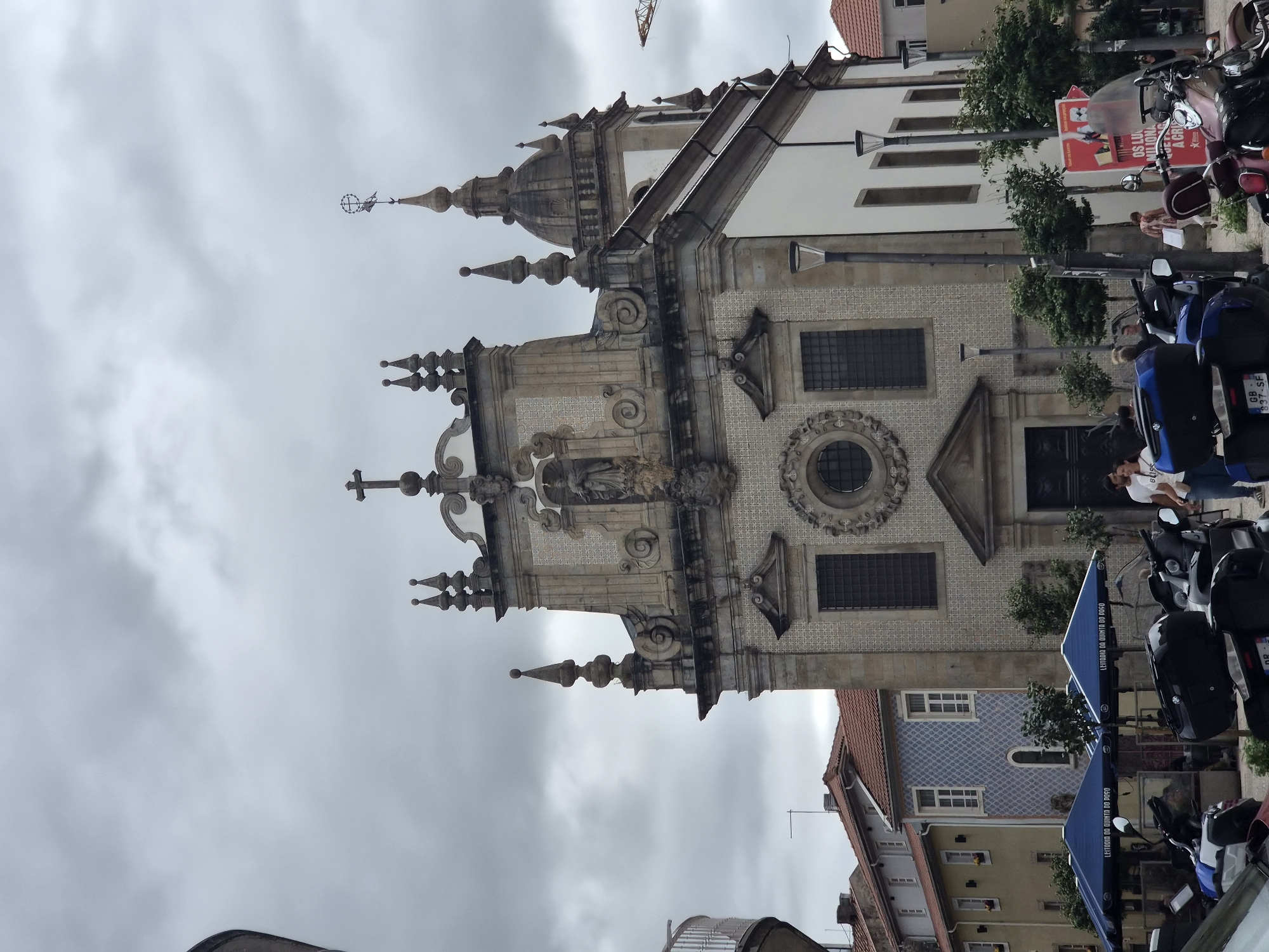 The image shows a view of the Church of Santa Maria Madalena in Braga, Portugal, under a cloudy sky. In the foreground, several motorcycles are parked, some covered with blue tarps. Blue umbrellas with text are also visible, suggesting an outdoor seating area. To the left, a spiral staircase leads up to a building. People are seen walking and standing near the entrance of the church, which has a highly ornate facade adorned with tiles and sculptures. The church features multiple spires and a dome. Adjacent to the church are buildings with tiled facades and traditional red-tiled roofs. A construction crane is visible in the distance.
