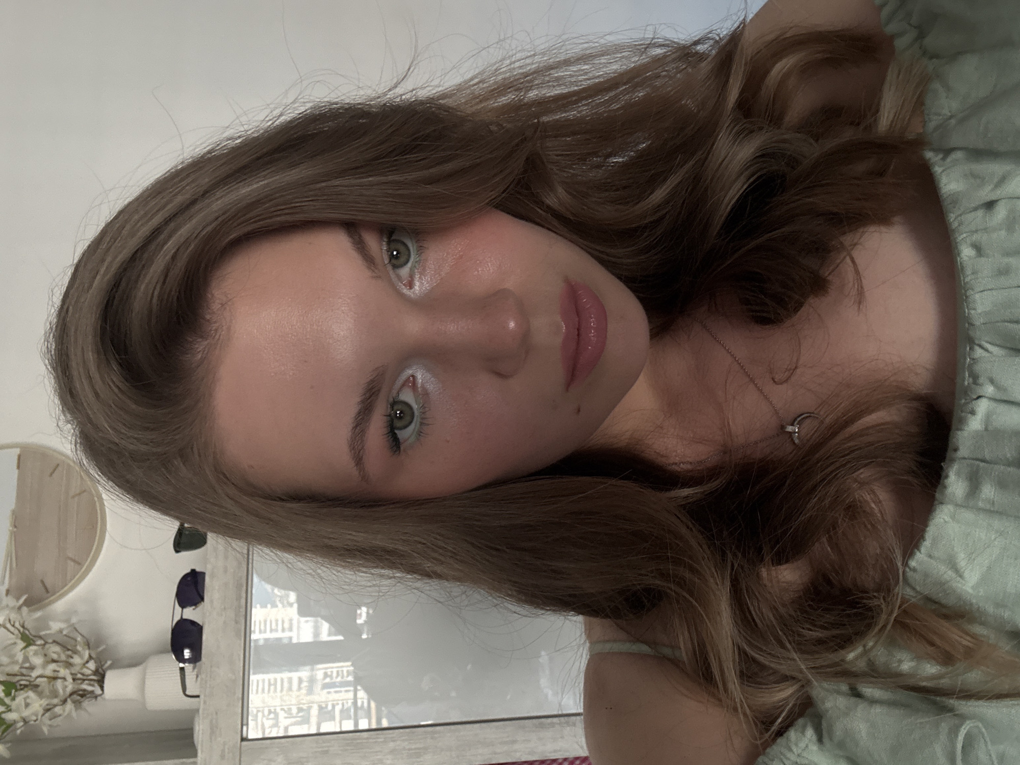 A young woman with long, wavy light brown hair and green eyes looks directly into the camera in a close-up, chest-up selfie. She has prominent white or light shimmery eyeshadow applied to the inner corners of her eyes and along the lower lash line, complemented by a thin line of light green eyeliner on her lower waterline. Her complexion displays a slight sheen, particularly on the forehead, cheekbones, and nose, indicative of highlight makeup. Her lips are full and covered in a glossy pink product. She wears a light green, off-the-shoulder top with a ruffled neckline and a thin silver necklace featuring a crescent moon pendant with a small embellishment. A small mole is visible on her right cheek. The background consists of a white wall. To her left, a mirror reflects an indoor setting with a white vase holding white artificial flowers and a round wooden decorative object on a shelf. Two pairs of sunglasses, one dark and one with green-tinted lenses, are also on the shelf. The mirror further reflects an urban landscape through a window, showing multiple modern buildings. This scene is situated indoors in the city of Budva, Montenegro.