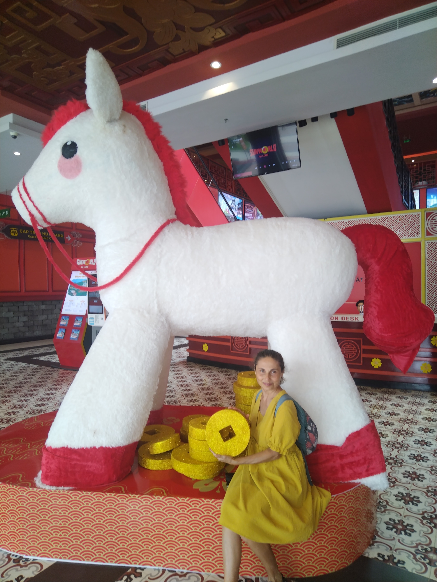 A woman poses next to a large plush horse in Bai Chay Ward, Vietnam. The horse is primarily white with red accents on its mane, tail, and hooves. The woman, wearing a yellow dress, sits on the base of the horse's display and holds a large, golden coin-shaped prop. The background features a red and gold decor, likely associated with a Chinese cultural theme. The location appears to be an indoor venue or establishment.